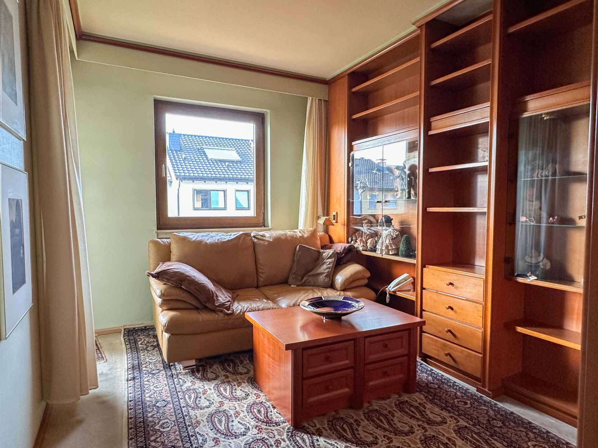 Wohnzimmer mit beiger Ledercouch, Holzregalwand, Teppich und Holztisch vor einem Fenster.
