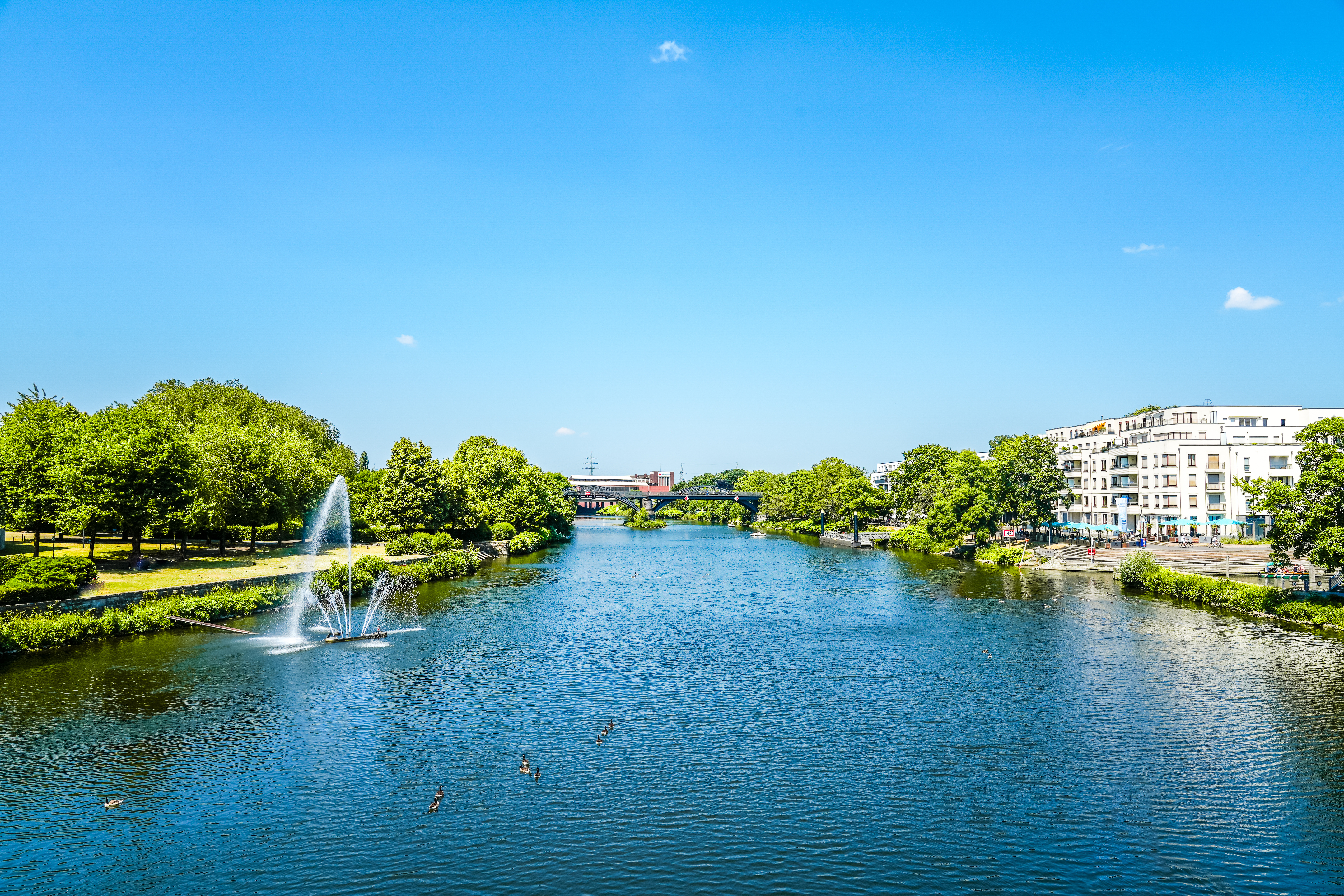 Fluss mit Springbrunnen, daneben Bäume und Wohnhäuser bei blauem Himmel.