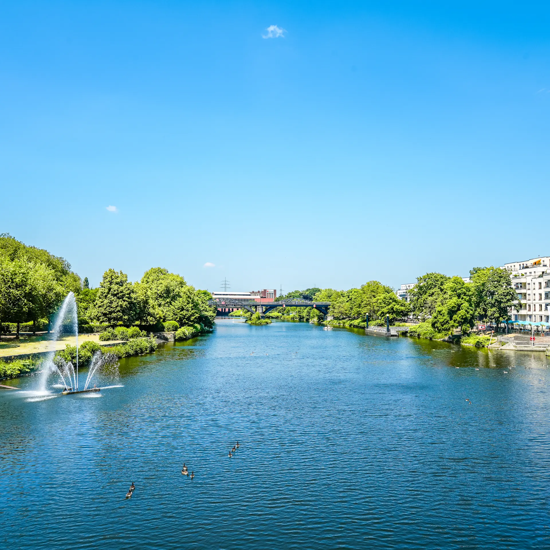 Fluss mit Springbrunnen, daneben Bäume und Wohnhäuser bei blauem Himmel.