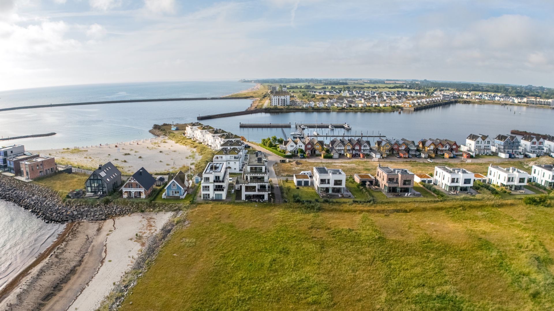 Luftaufnahme von modernen Wohnhäusern an einem Sandstrand und einem kleinen Hafen.