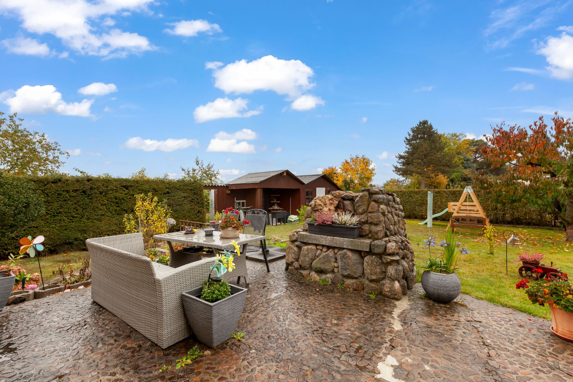 Garten mit Terrasse, Steingrill, Sitzgruppe, Pflanzen, Spielgerüst und Gartenhaus bei blauem Himmel.