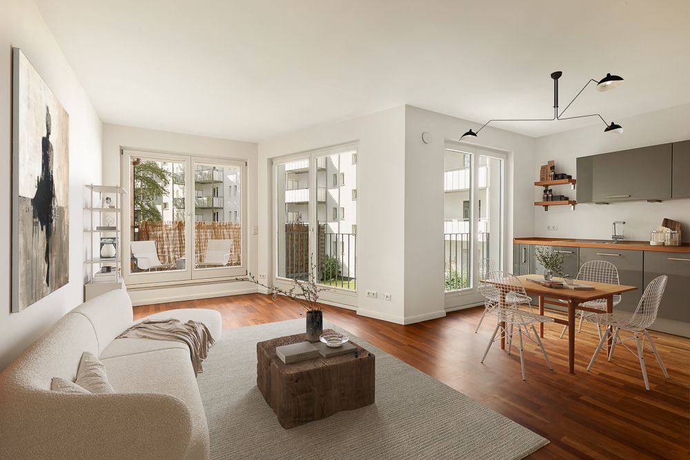 Helles Wohnzimmer mit Sofa, Couchtisch, Esstisch und offener Küche, große Fenster und Holzboden.