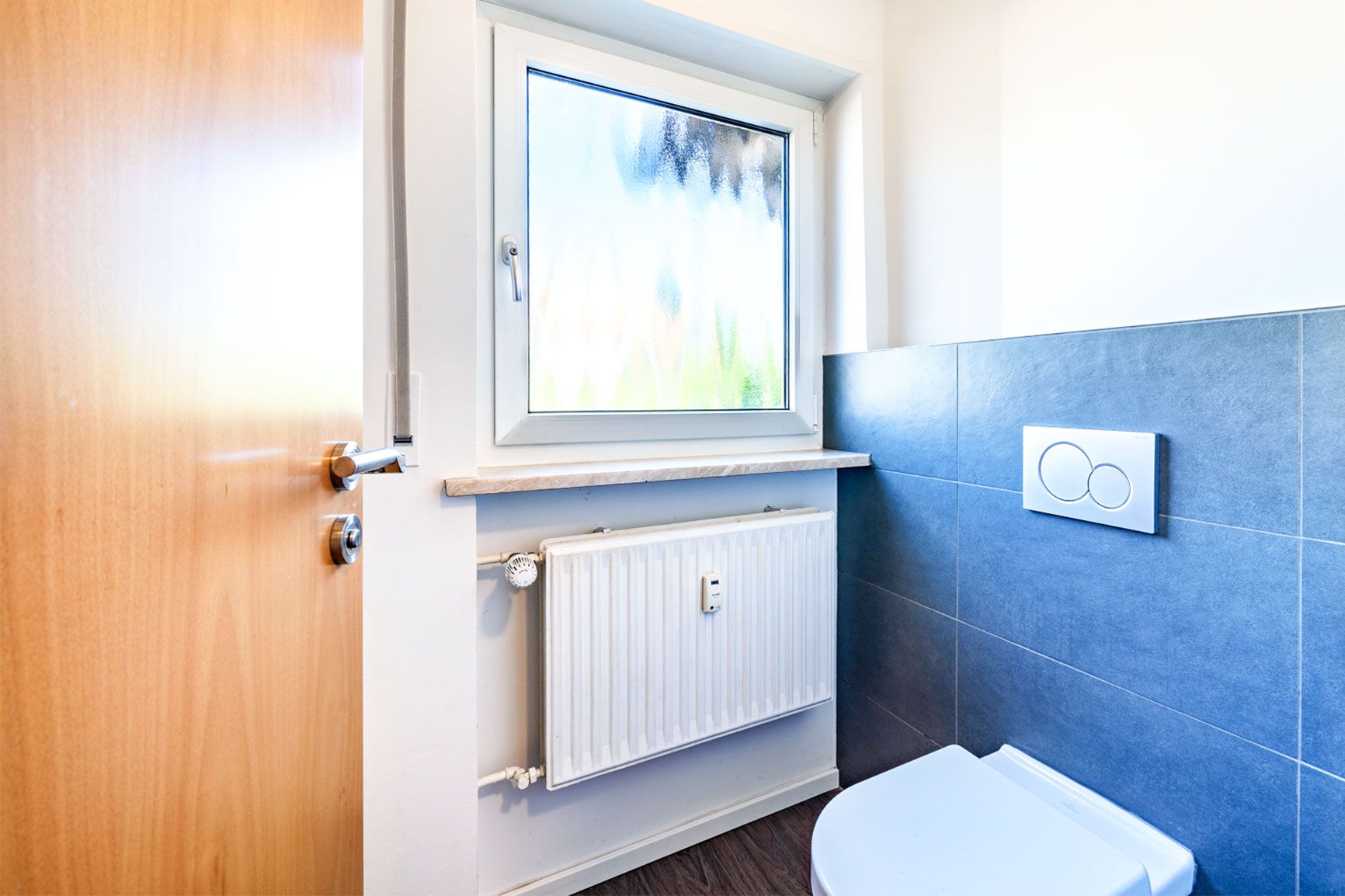 Kleines Badezimmer mit Toilette, Fenster und Heizkörper, blaue Wandfliesen.