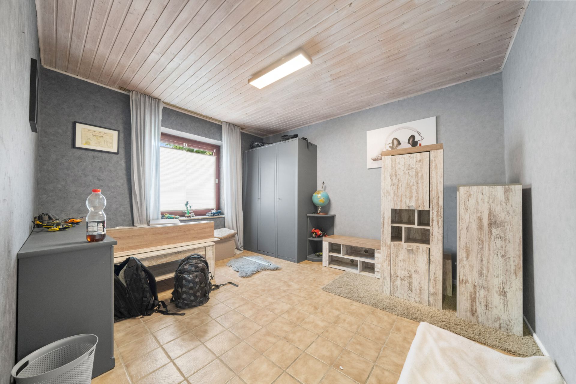 Helles Zimmer mit Schrank, Kommode, zwei Rucksäcken und einem Fenster mit Vorhängen.