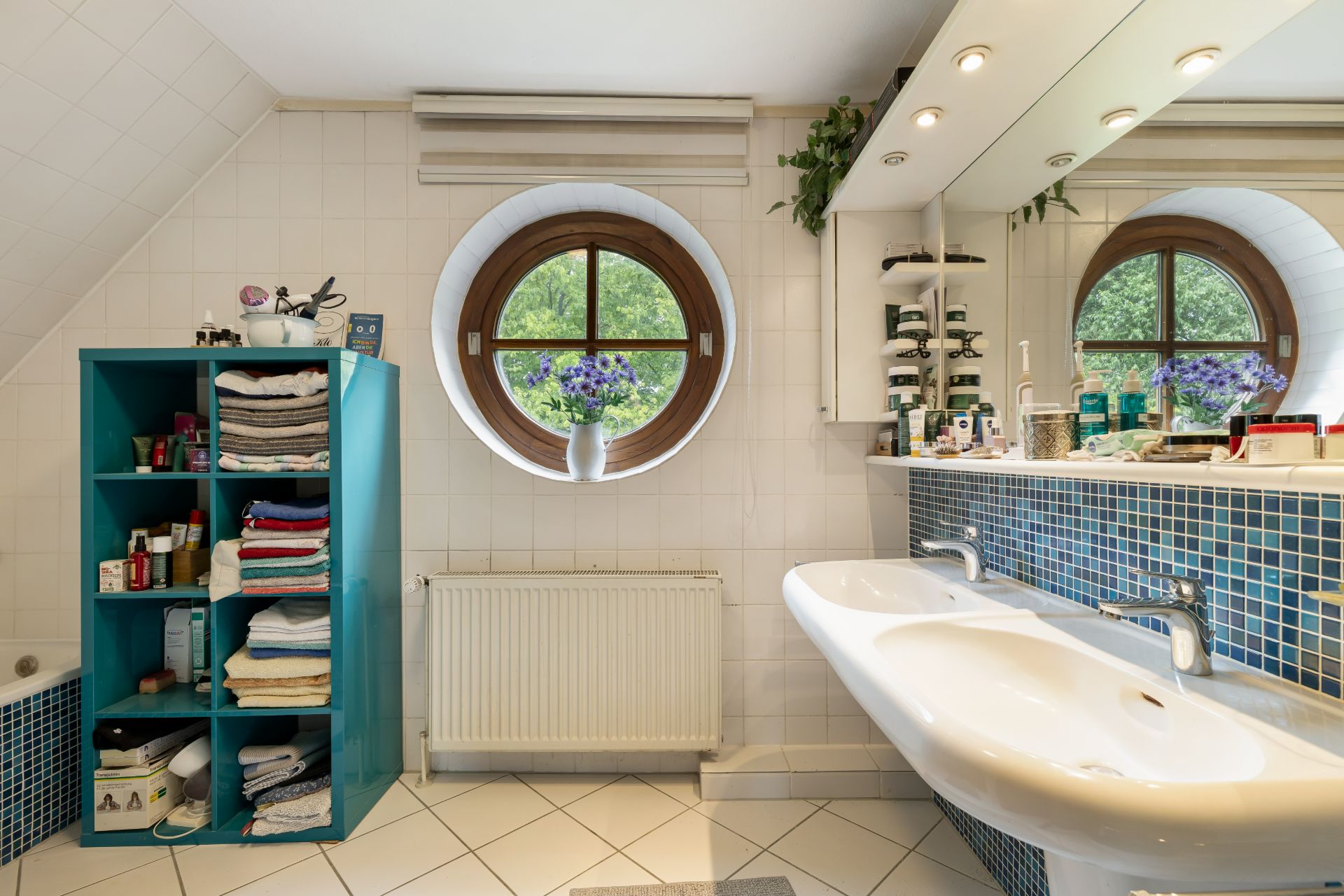 Modernes Badezimmer mit rundem Fenster, Doppelwaschbecken und blauem Regal mit Handtüchern.