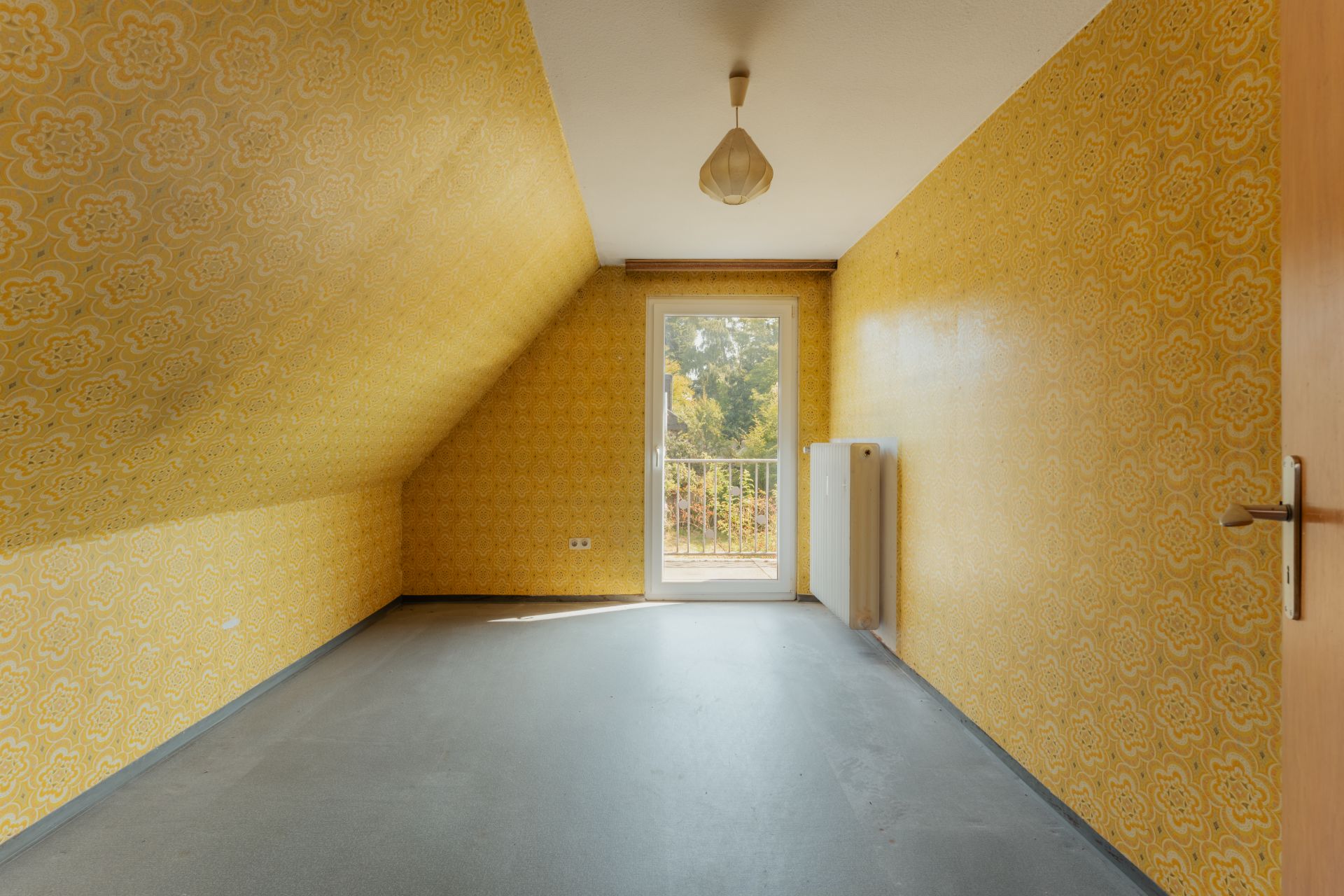 Leeres Zimmer mit gelber Tapete, grauem Boden und Tür zum Balkon.