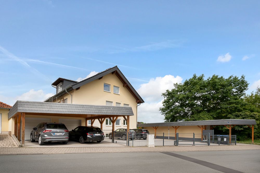 Wohnhaus mit Carport und mehreren geparkten Autos, im Hintergrund Bäume und blauer Himmel.