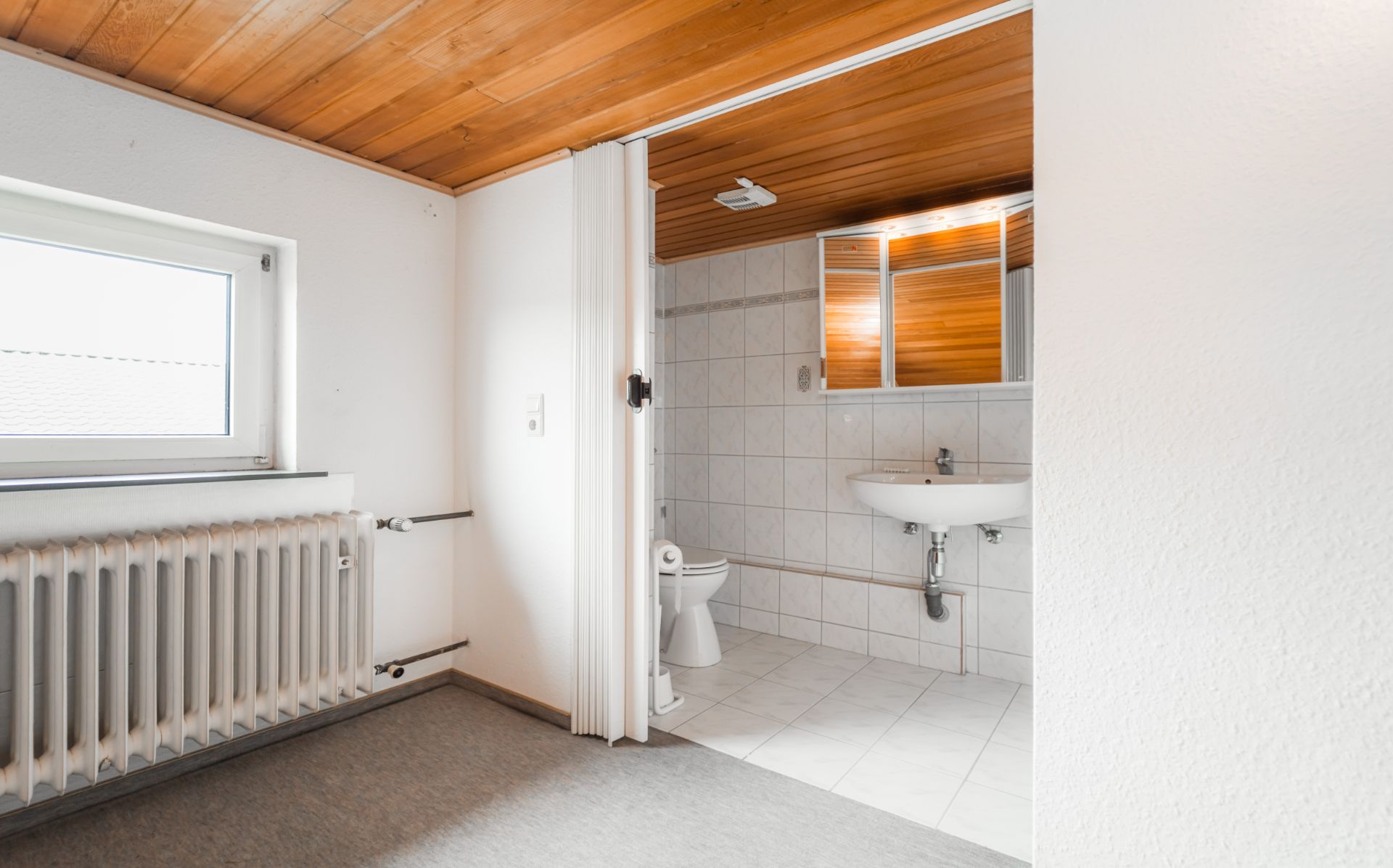 Helles Badezimmer mit Waschbecken, WC, Spiegel und Holzdecke, Blick vom benachbarten Raum.