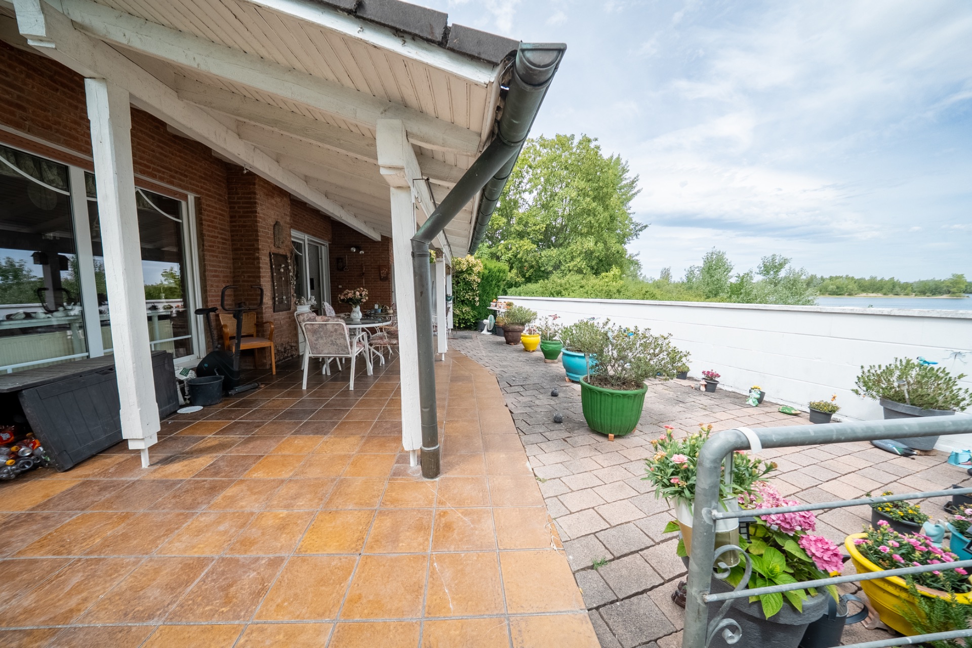 Überdachte Terrasse mit braunen Fliesen, weißen Möbeln und vielen bunten Blumentöpfen.