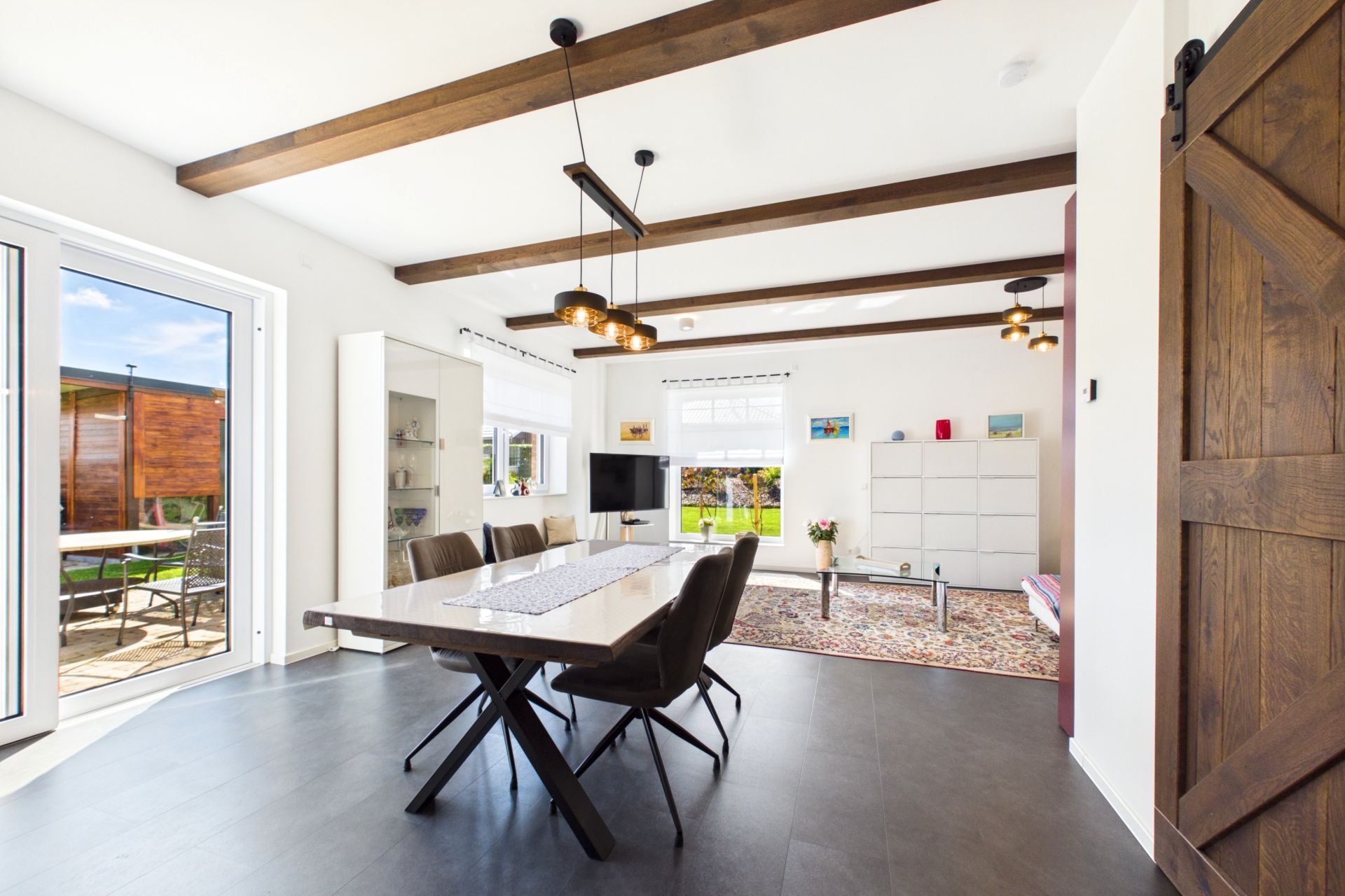 Modernes Ess- und Wohnzimmer mit Holzbalken, großem Tisch, Sofa und Zugang zur Terrasse.