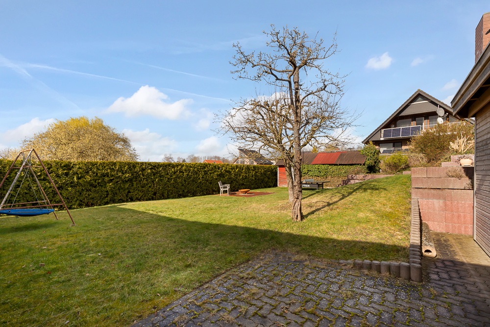 Grüner Garten mit Baum, Schaukel, Sitzbank und gepflastertem Bereich bei Wohnhaus.