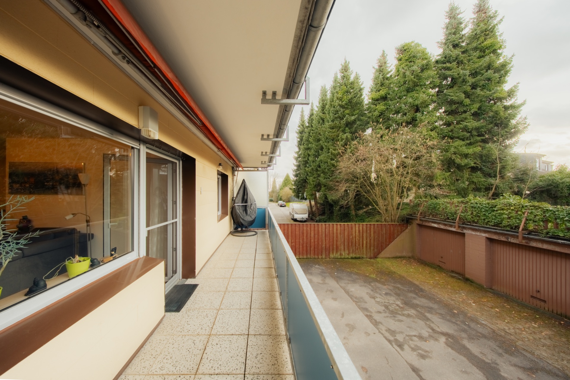 Langer Balkon mit Glasgeländer, Blick auf Garten und Einfahrt, Bäume im Hintergrund.