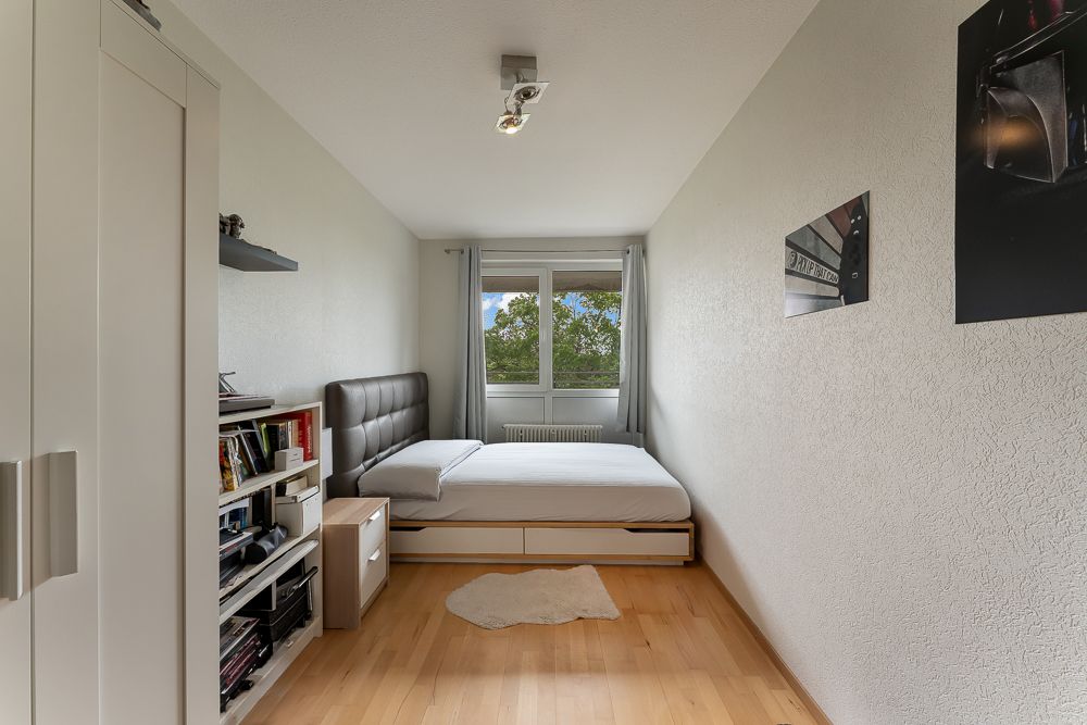 Schlafzimmer mit Bett, Bücherregal, Nachttisch und großem Fenster mit Blick auf Bäume.