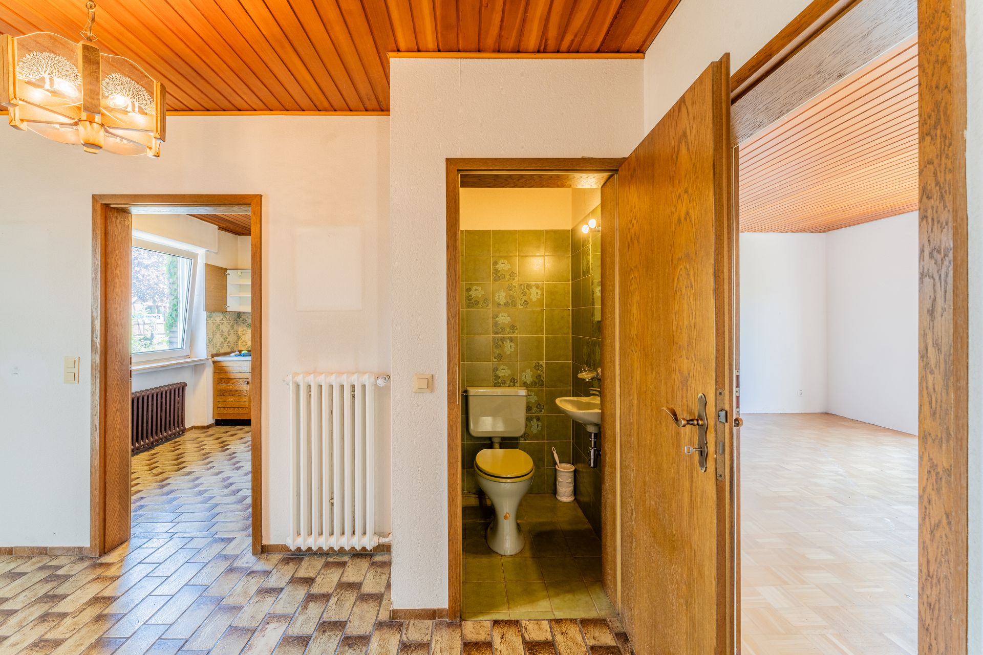 Innenraum mit Flur, WC, Küche und angrenzendem Wohnzimmer. Holzdecke und Fliesenboden sichtbar.