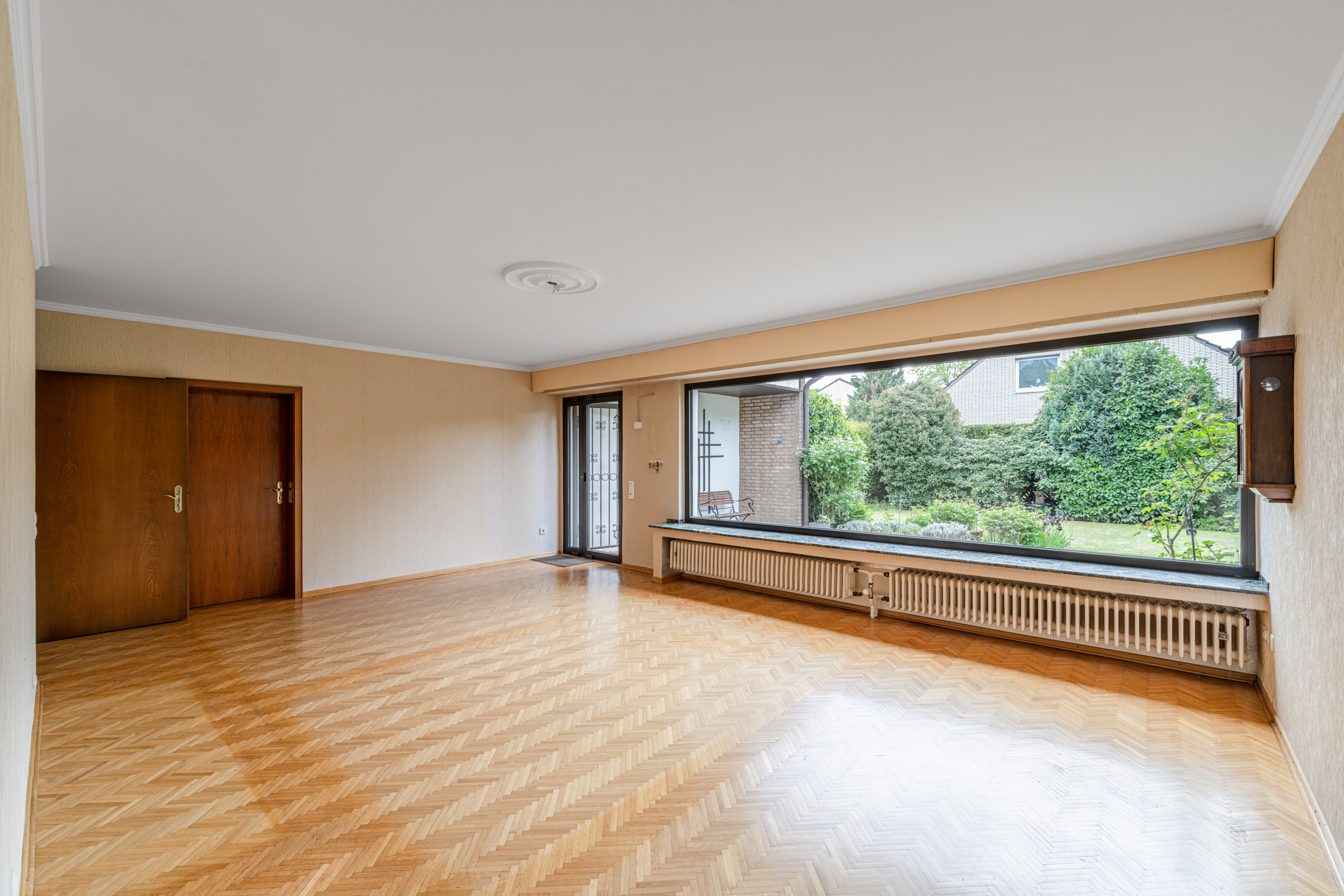 Leeres Wohnzimmer mit Parkettboden und großem Fenster mit Blick in den Garten.