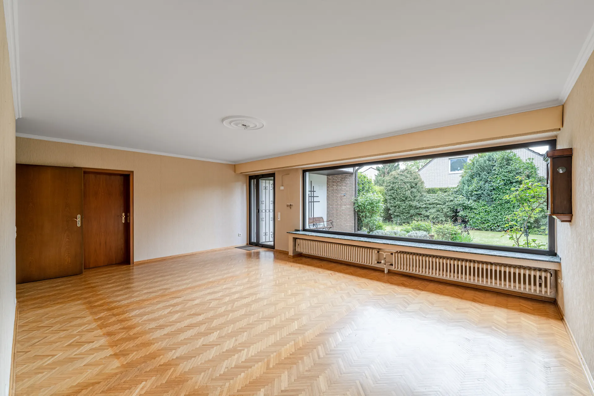 Leeres Wohnzimmer mit Parkettboden und großem Fenster mit Blick in den Garten.