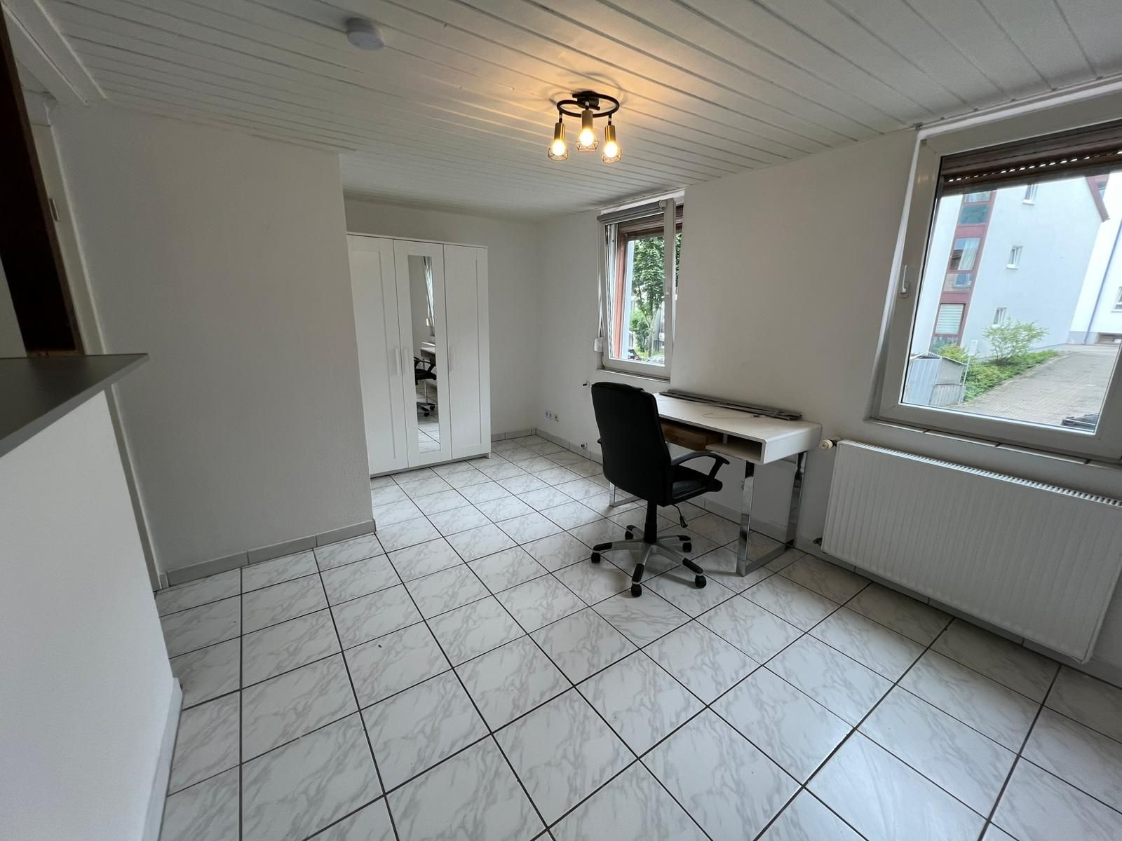 Helles Zimmer mit Fliesenboden, Schreibtisch, Bürostuhl, Kleiderschrank und zwei Fenstern.