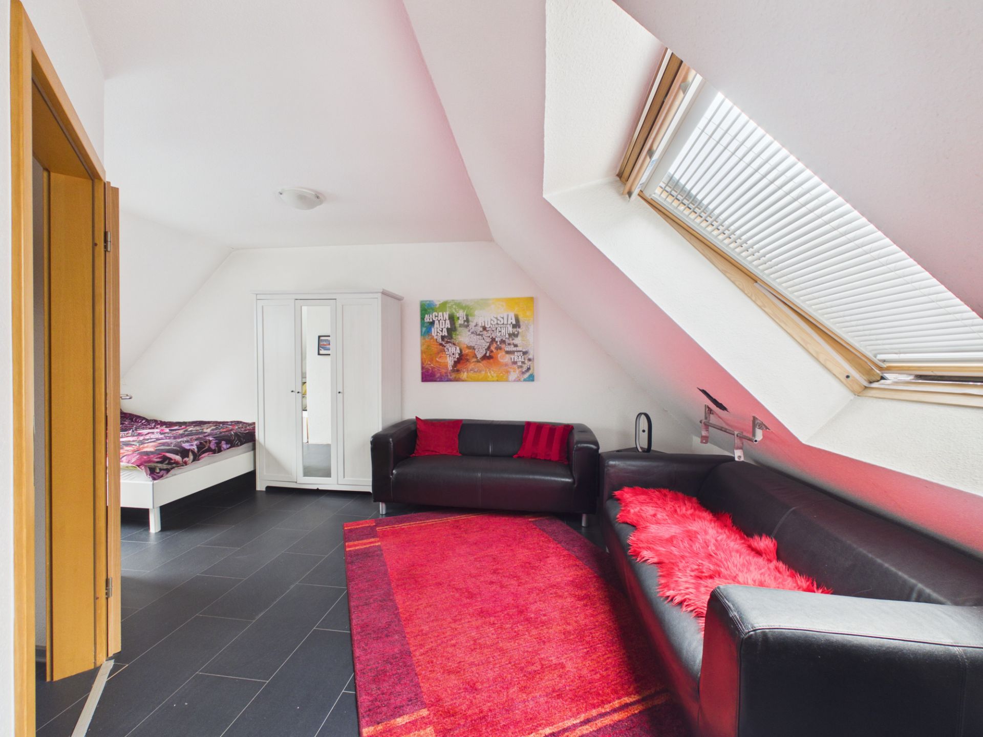 Helles Dachzimmer mit Sofa, Teppich, Bett, Kleiderschrank und schrägem Fenster.
