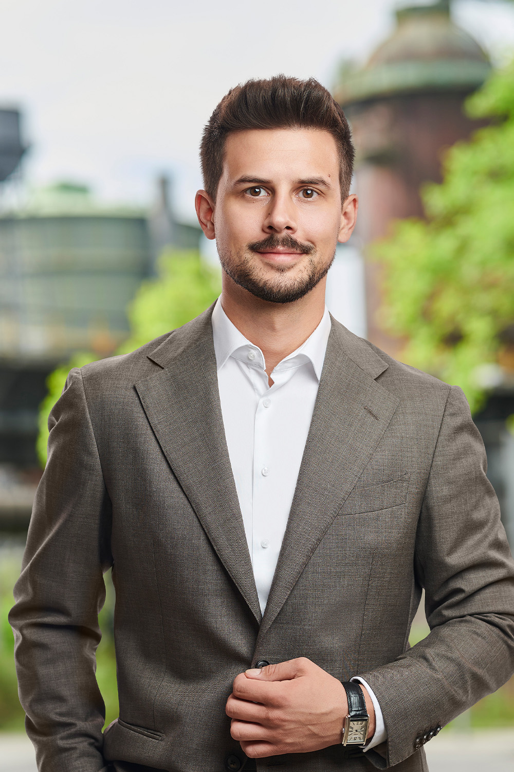 Ein Mann im Anzug steht draußen vor unscharfem Hintergrund mit Gebäuden und Bäumen.