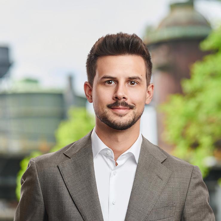 Ein Mann im Anzug steht draußen vor unscharfem Hintergrund mit Gebäuden und Bäumen.