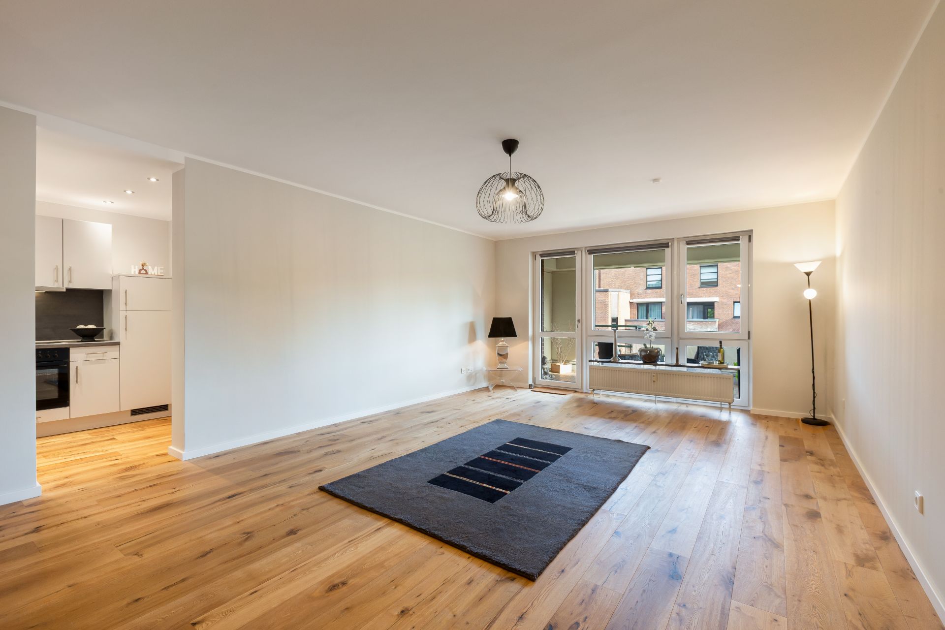 Helles Wohnzimmer mit Holzfußboden, Teppich, Stehlampe und Zugang zum Balkon, offene Küche links.
