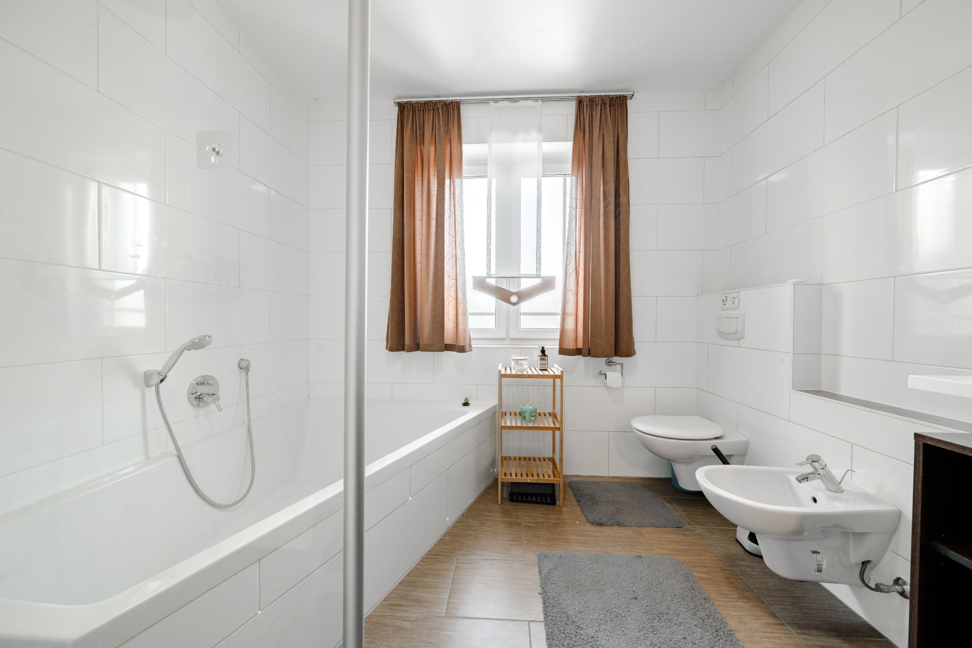 Modernes Badezimmer mit Badewanne, Waschbecken, Bidet und Fenster mit braunen Vorhängen.