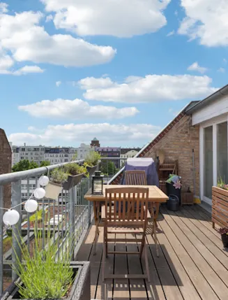 Holzbalkon mit Holztisch, Stühlen, Pflanzen und Lichterkette an einem Wohnhaus.