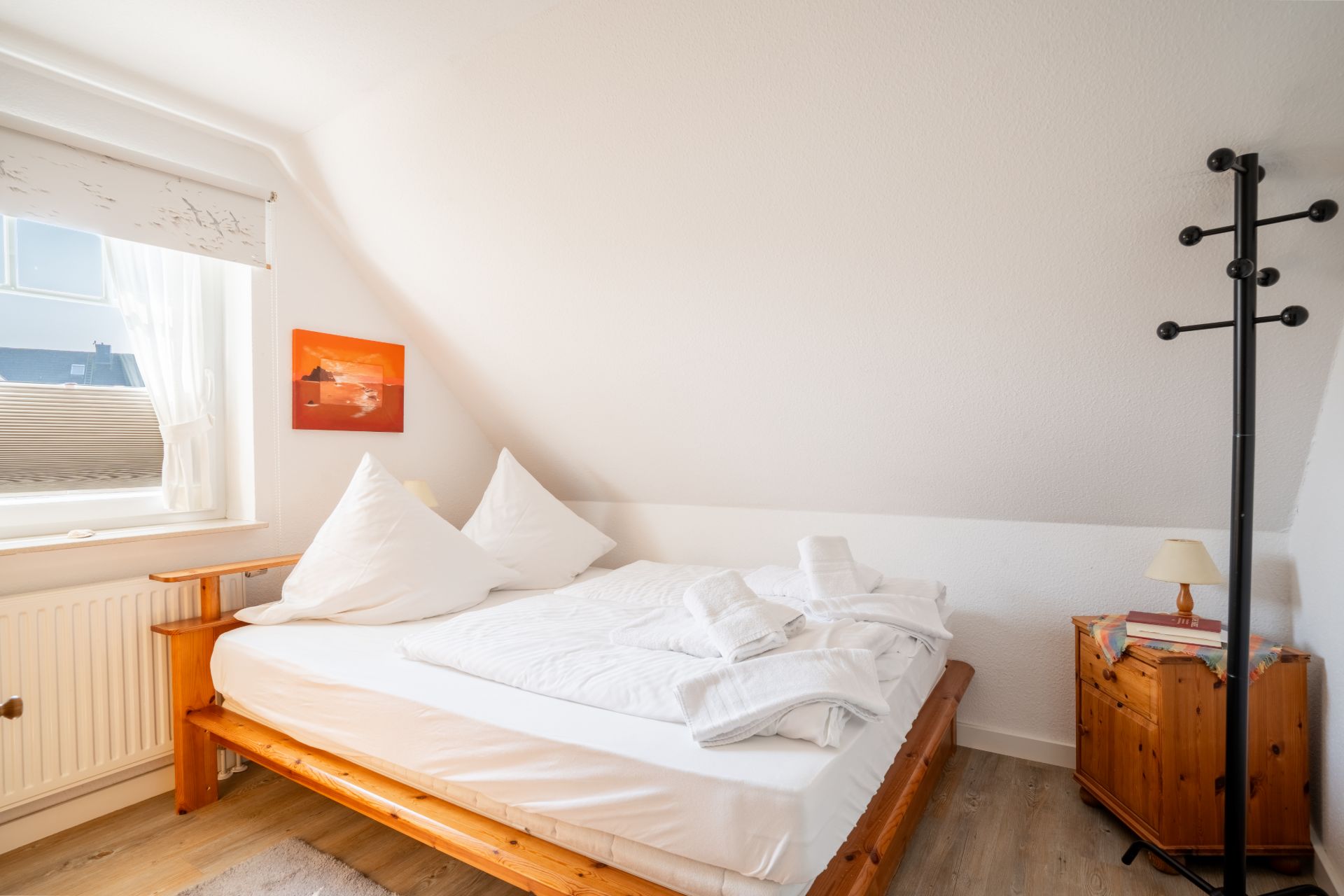 Schlafzimmer mit Holz-Doppelbett, Nachttisch, Kleiderständer und Fenster mit Vorhang.