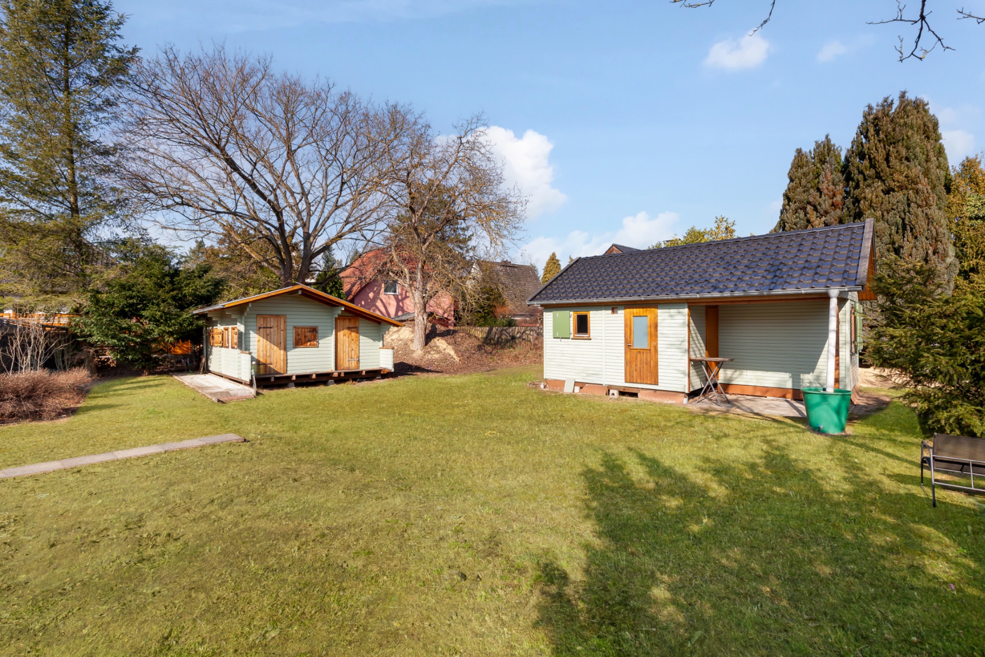 Zwei kleine Gartenhäuser stehen auf einer großen, grünen Rasenfläche mit Bäumen und Sträuchern.