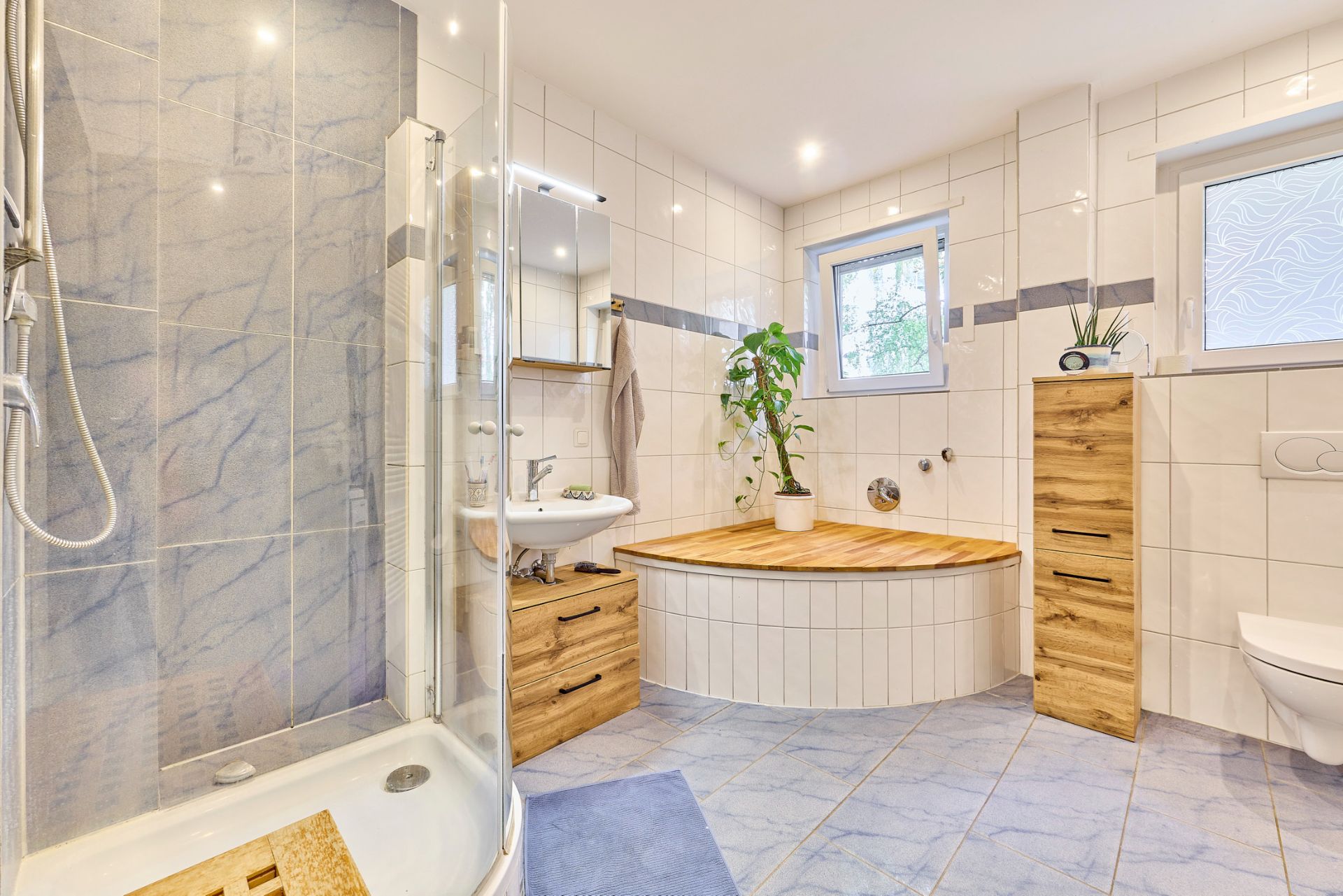 Modernes Badezimmer mit Dusche, Eckbadewanne, Holzschränken und zwei Fenstern.