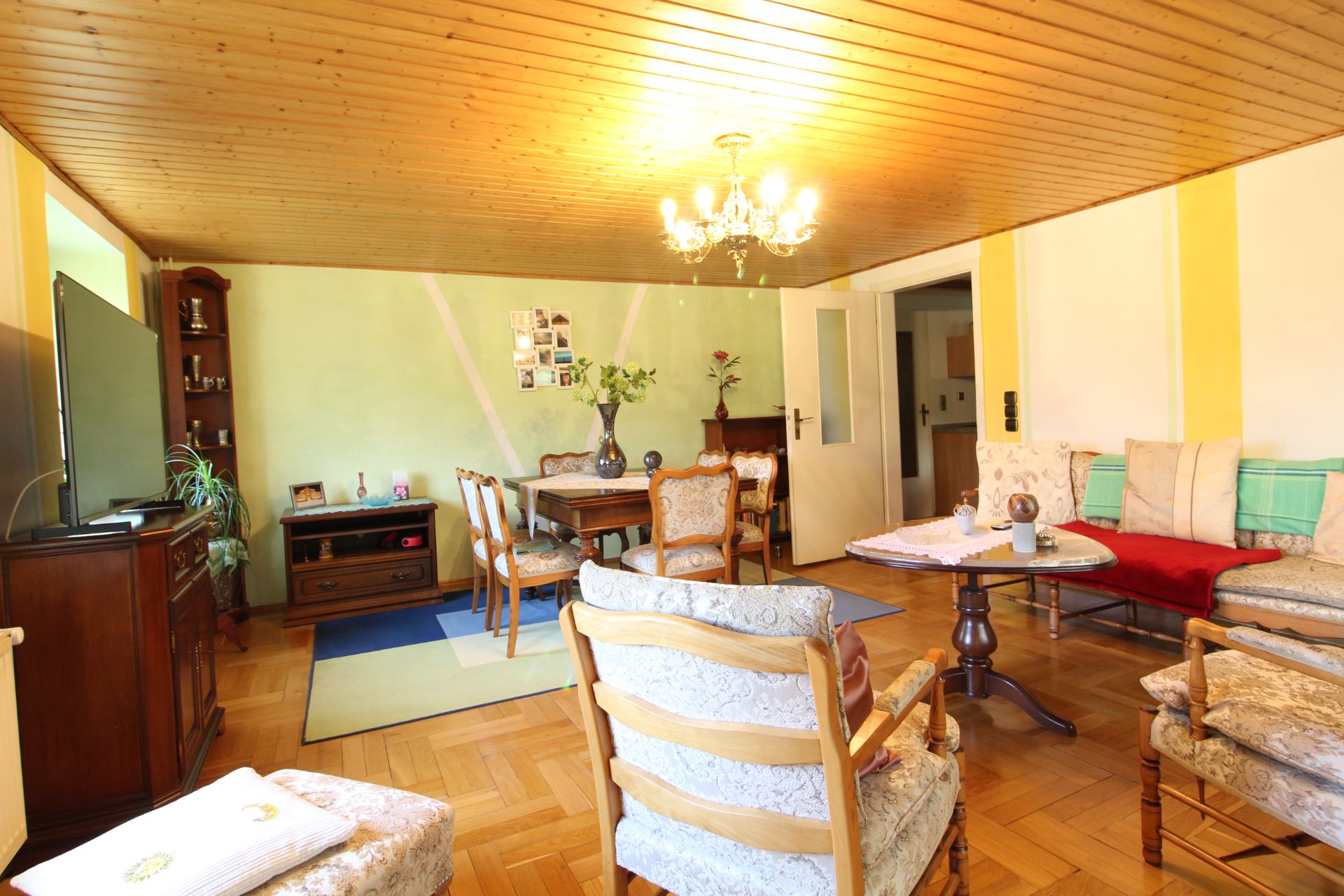 Helles Wohnzimmer mit Holzboden, Sitzgruppe, Esstisch, TV und Kronleuchter.