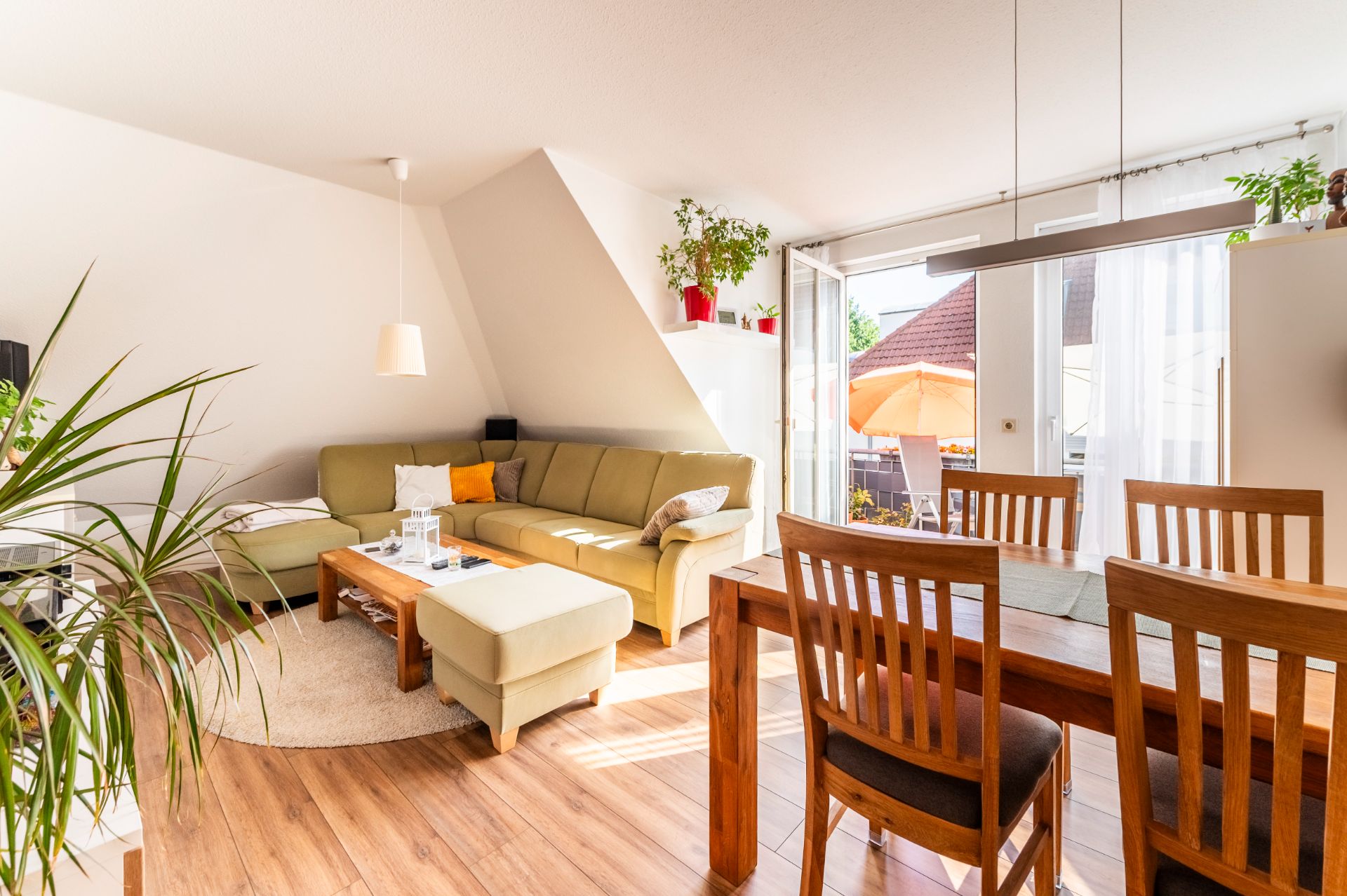 Helles Wohnzimmer mit Ecksofa, Holztisch, Esstisch und Stühlen, Pflanzen und Zugang zum Balkon.