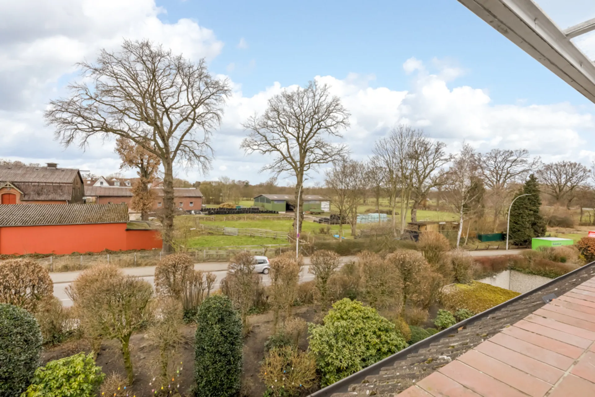 Blick von einer Terrasse auf eine Straße, Bäume und ländliche Gebäude mit Wiese.