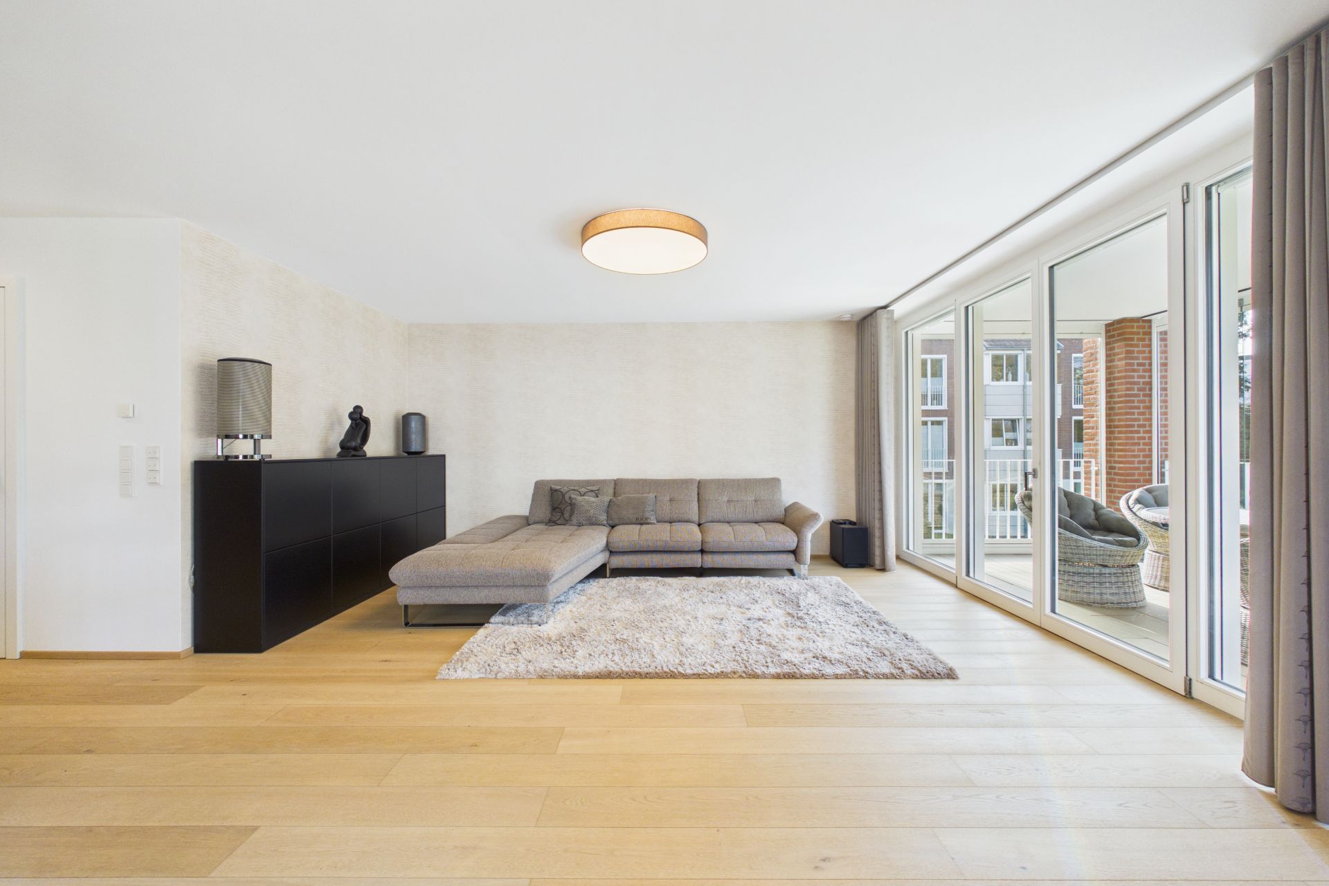Helles Wohnzimmer mit grauem Sofa, Teppich, Sideboard und großen Fenstern zur Terrasse.