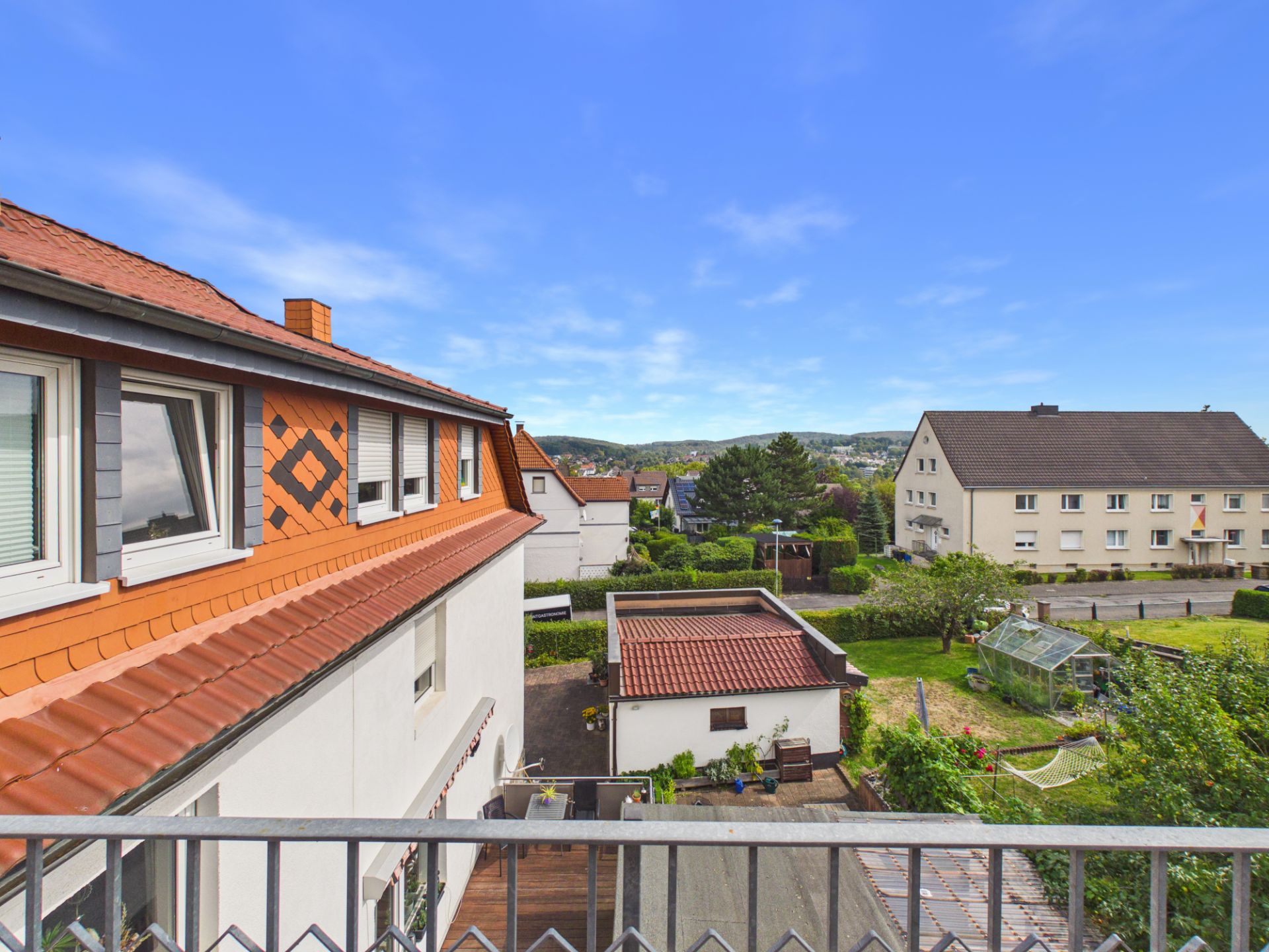Blick von einem Balkon auf Nachbarhäuser, Garten und Gewächshaus bei klarem Himmel.