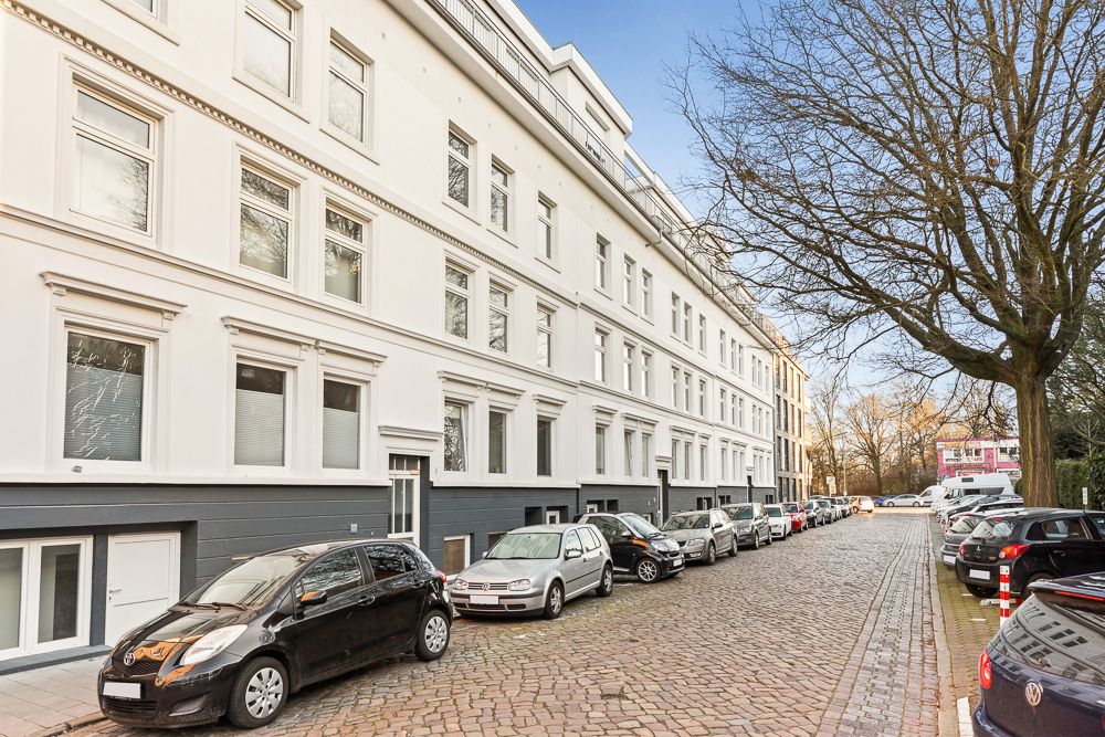 Wohnhaus mit vielen Fenstern an einer Kopfsteinpflasterstraße, geparkte Autos am Straßenrand.
