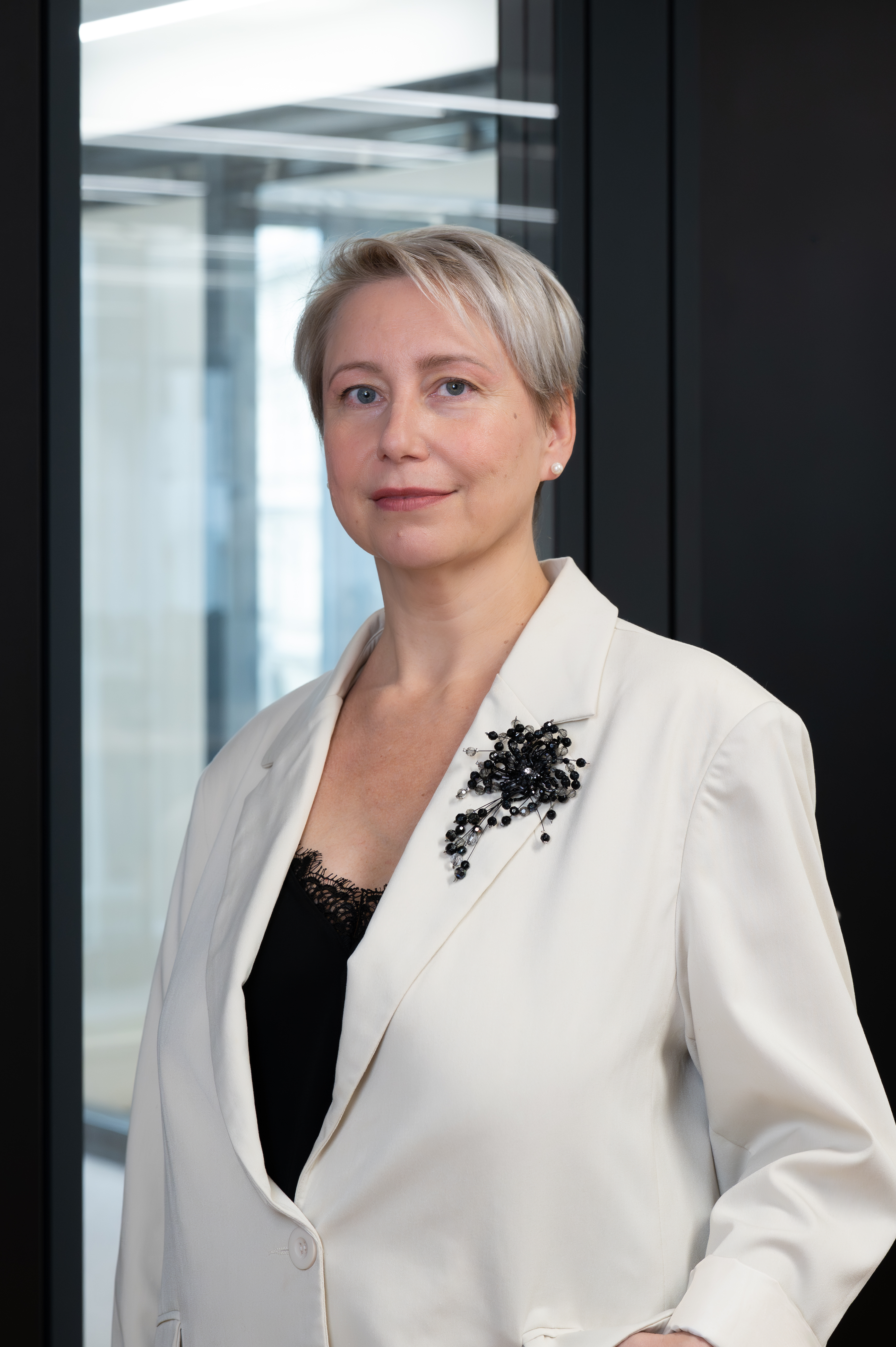 Frau mit kurzem, blondem Haar im hellen Blazer steht vor einem Fenster.