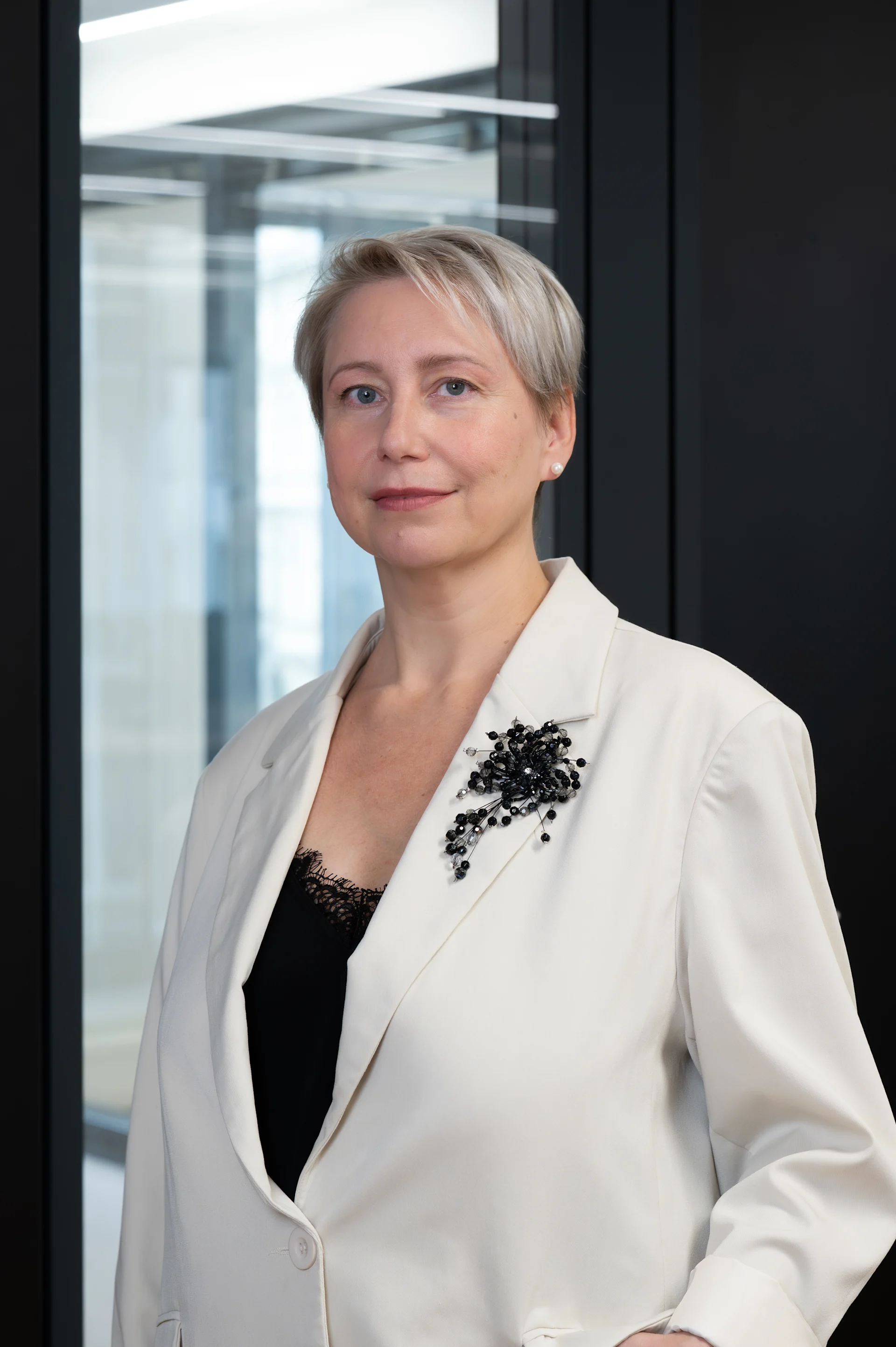 Frau mit kurzem, blondem Haar im hellen Blazer steht vor einem Fenster.