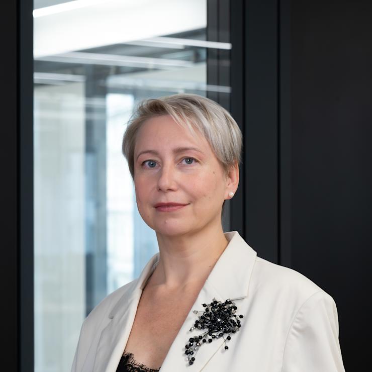 Frau mit kurzem, blondem Haar im hellen Blazer steht vor einem Fenster.