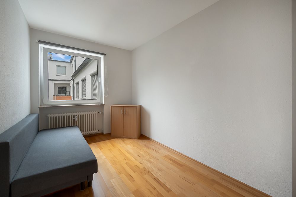 Kleiner Raum mit grauem Sofa, Fenster, Heizkörper und Holzschrank auf Parkettboden.