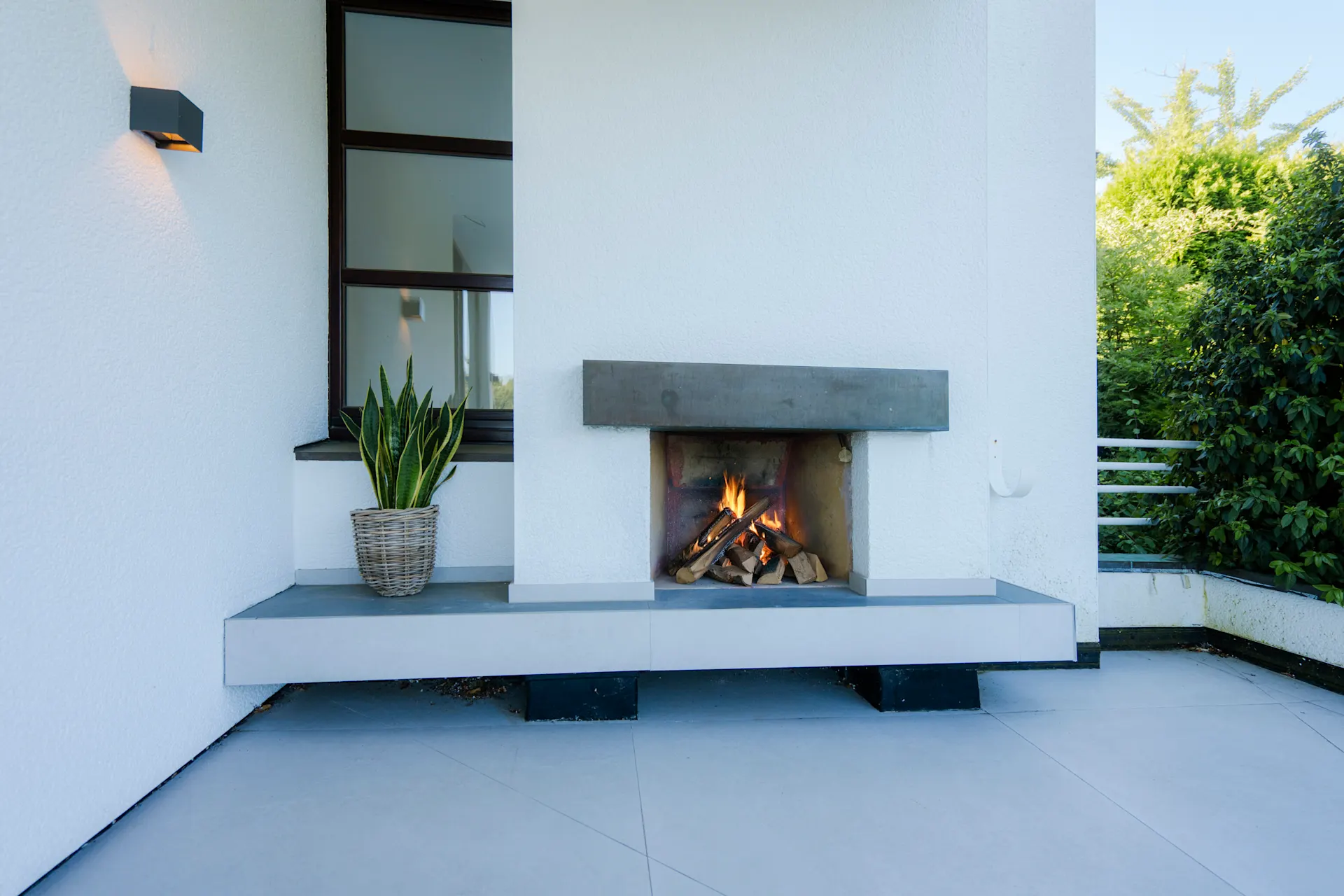 Moderne Terrasse mit offenem Kamin, Pflanze im Korb und Fenster im Hintergrund.