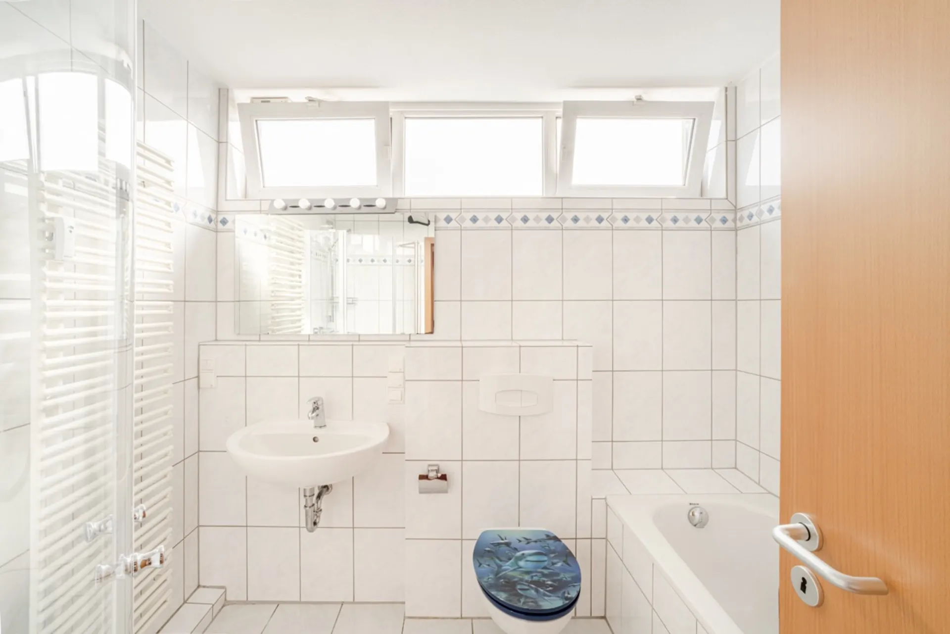 Helles Badezimmer mit weißer Fliesen, Waschbecken, Toilette, Badewanne und Fenster.