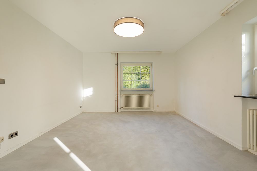 Leerer, heller Raum mit Fenster, Heizung und Deckenlampe, Wände und Boden sind hell.