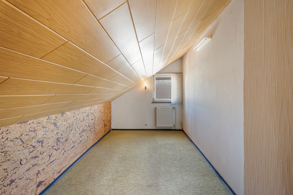 Langer Raum mit schräger Holzdecke, Fenster und Heizkörper am Ende.