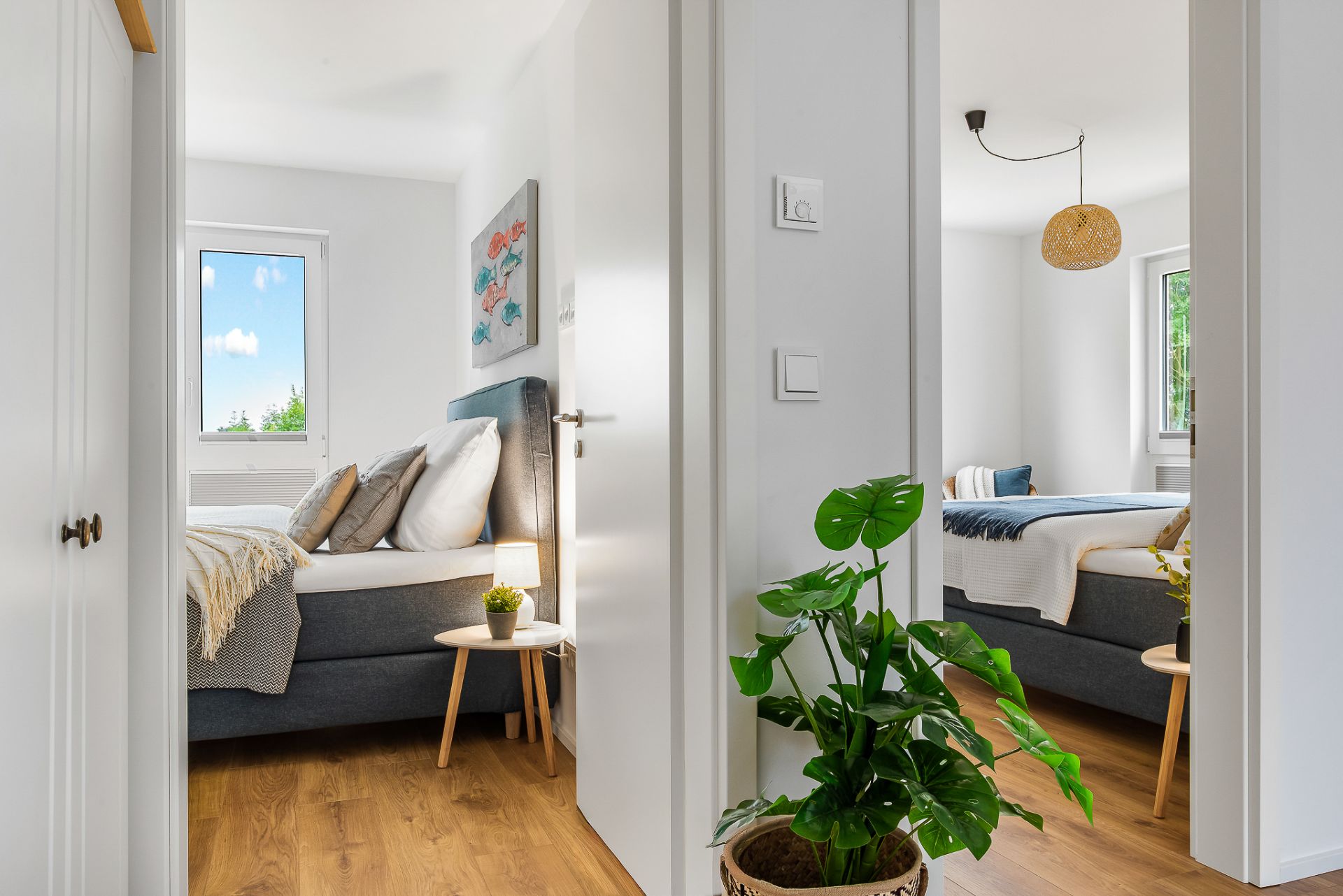 Blick in zwei helle Schlafzimmer mit Doppelbetten und Holzböden, dazwischen eine grüne Zimmerpflanze.