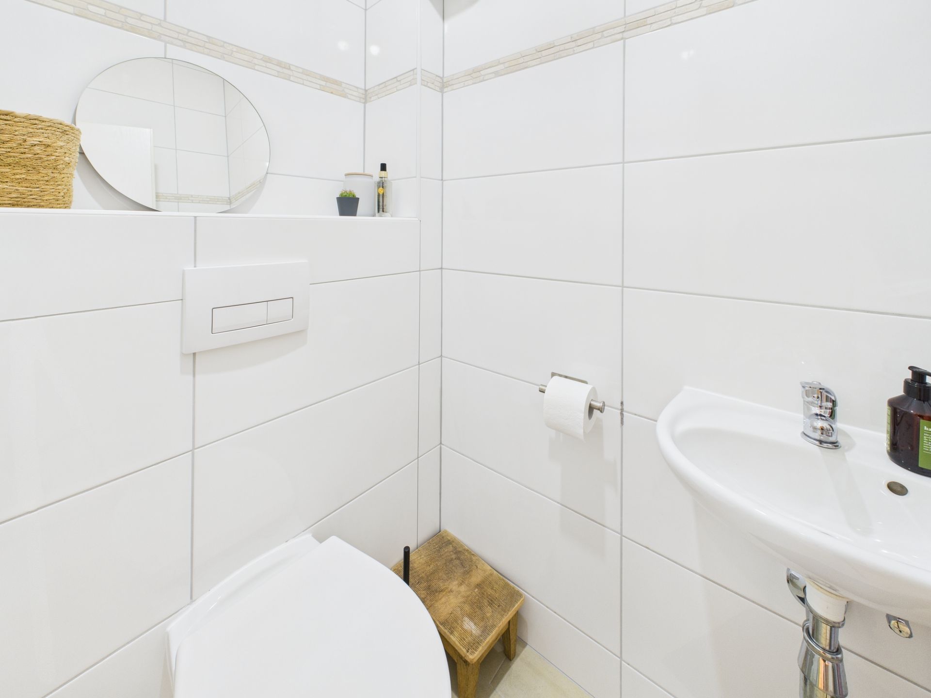 Modernes, weiß gefliestes Badezimmer mit Toilette, Wandwaschbecken und kleinem Spiegel.