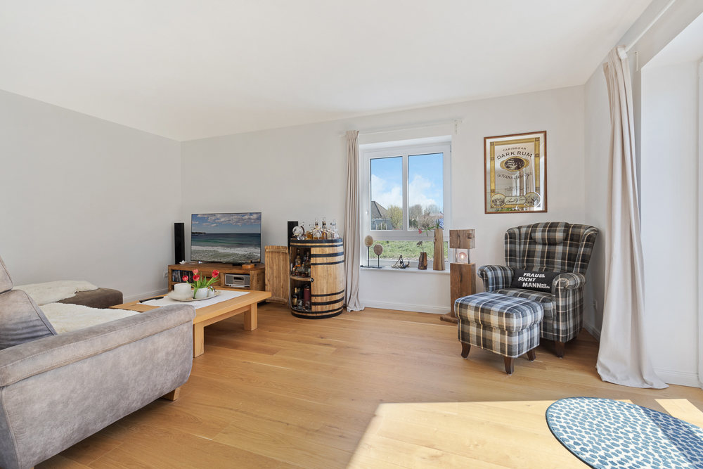 Helles Wohnzimmer mit Sofa, Couchtisch, Fernseher, gemustertem Sessel und Fenster mit Blick nach draußen.
