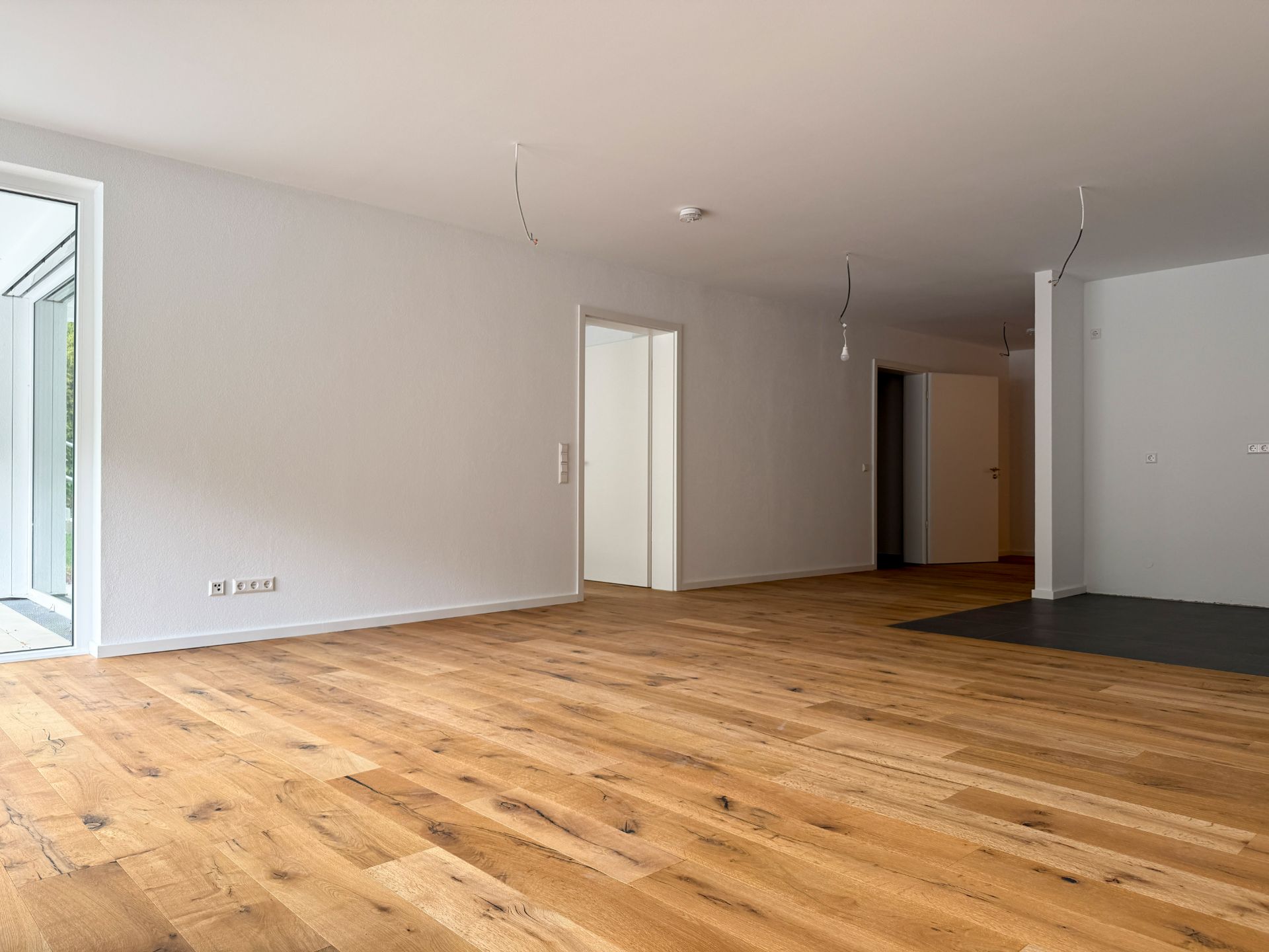 Leeres Wohnzimmer mit Holzfußboden und weißen Wänden, Türen führen in andere Räume.