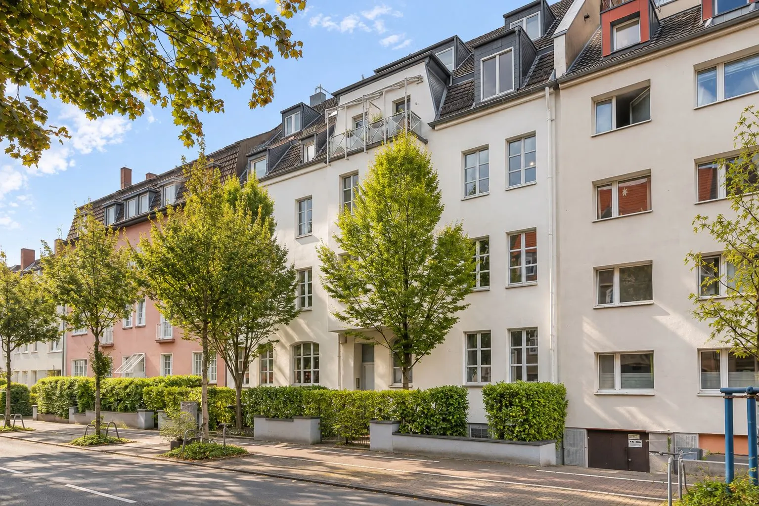 Mehrstöckiges Wohngebäude mit Bäumen und Sträuchern vor dem Eingang an einer ruhigen Straße.