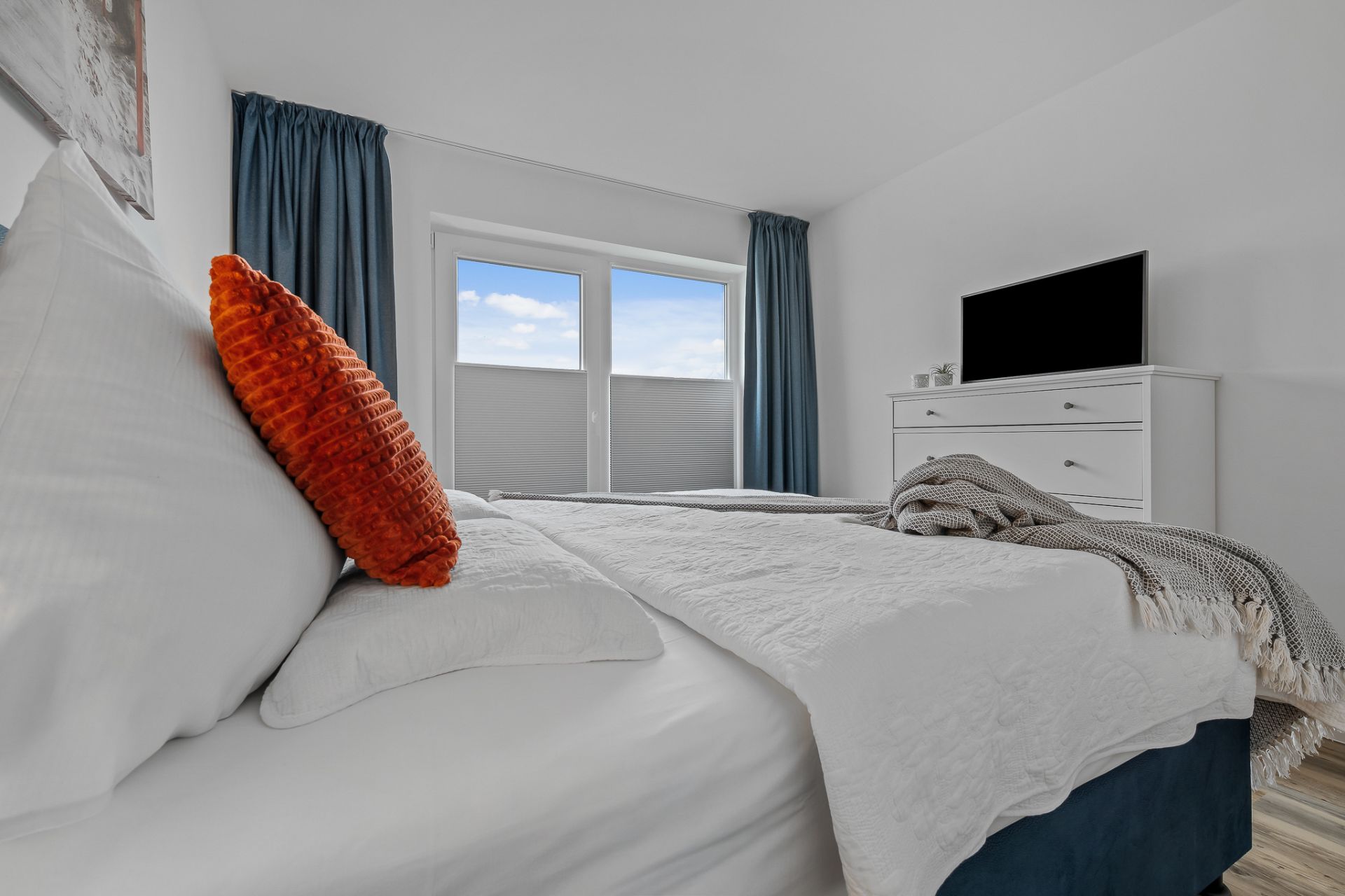Schlafzimmer mit großem Bett, orange Kissen, Kommode mit Fernseher und Fenster mit blauen Vorhängen.